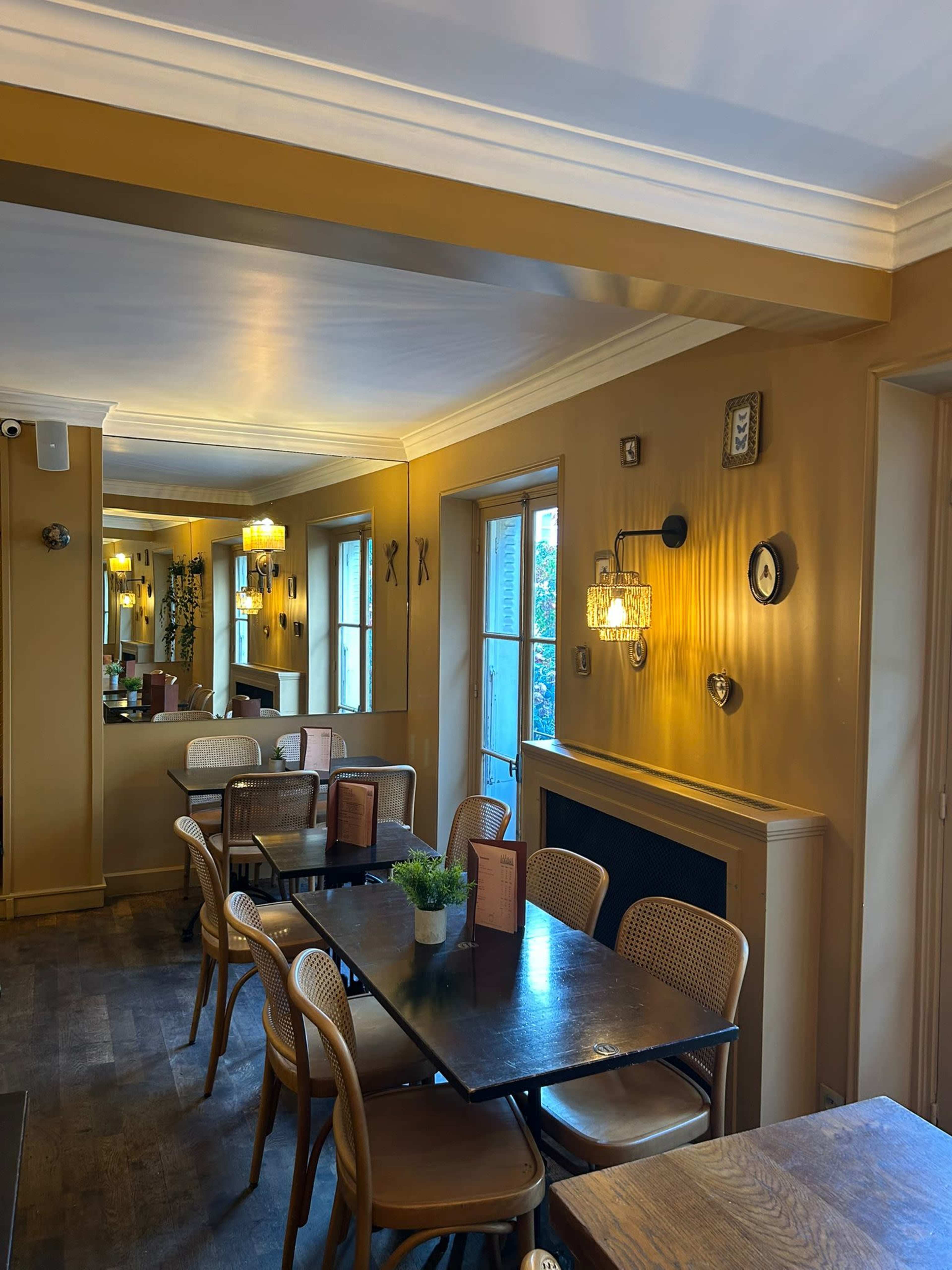 The image shows a cozy restaurant interior featuring wooden tables, woven chairs, and warm golden walls with framed decor.