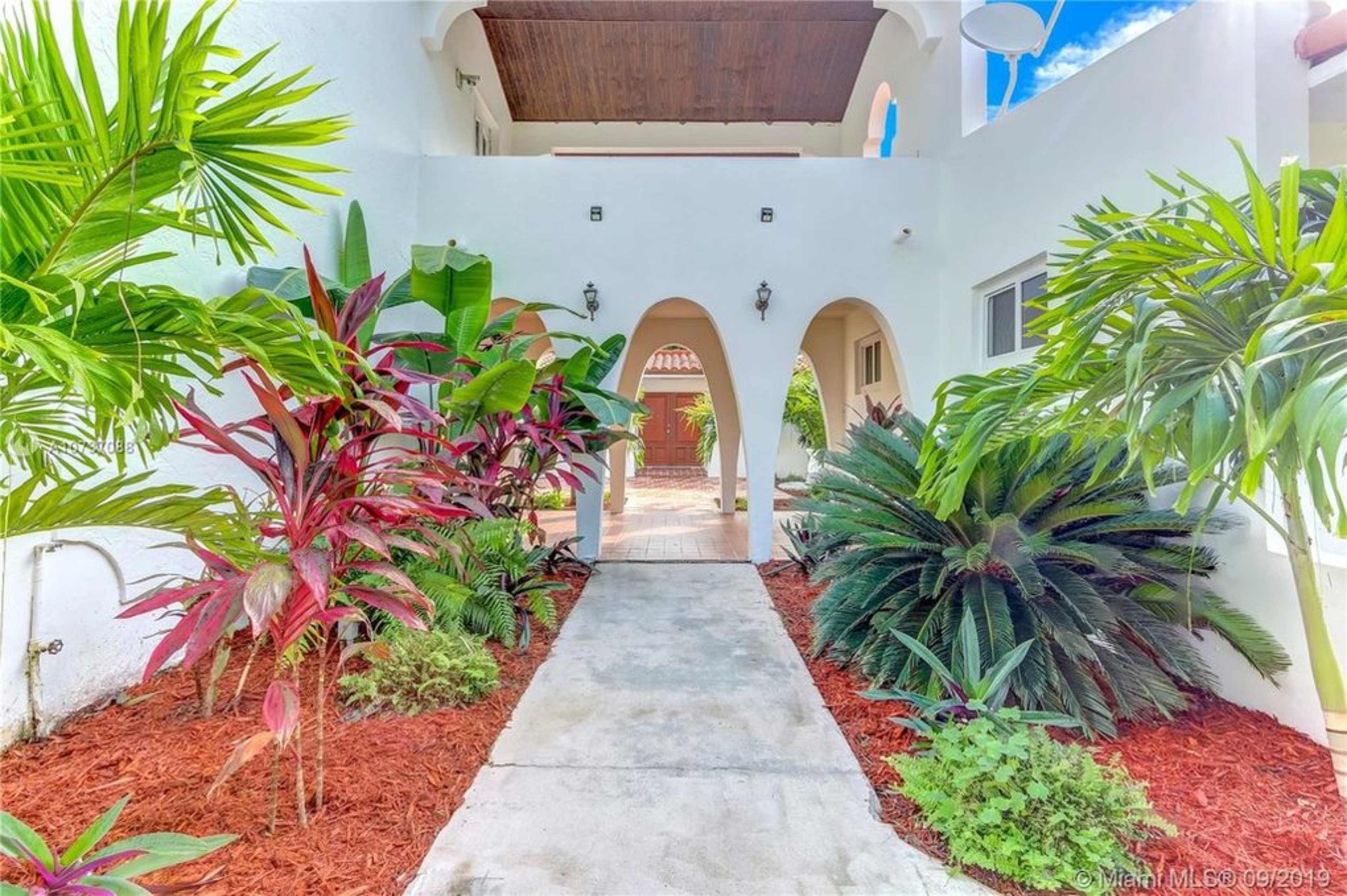A pathway leads through a landscaped entrance with tropical plants and an arched entryway to a courtyard.