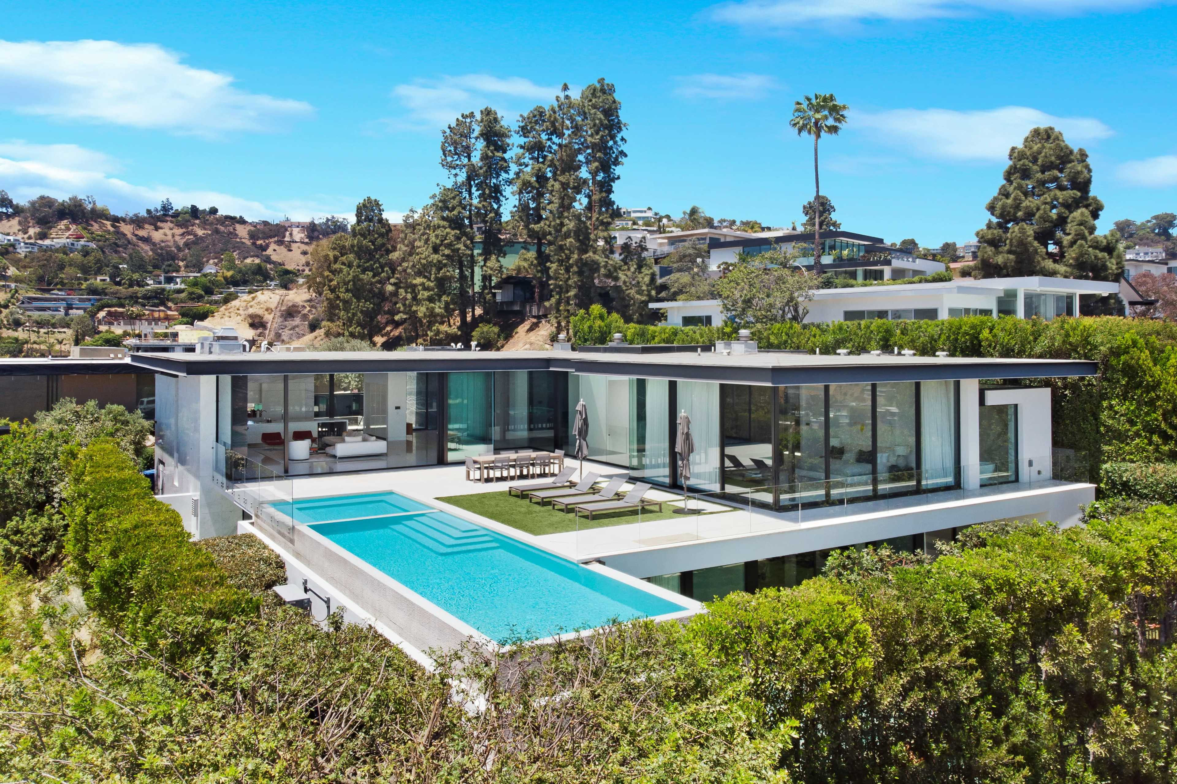 The image shows a modern glass-fronted house with a swimming pool surrounded by greenery, located on a hillside.