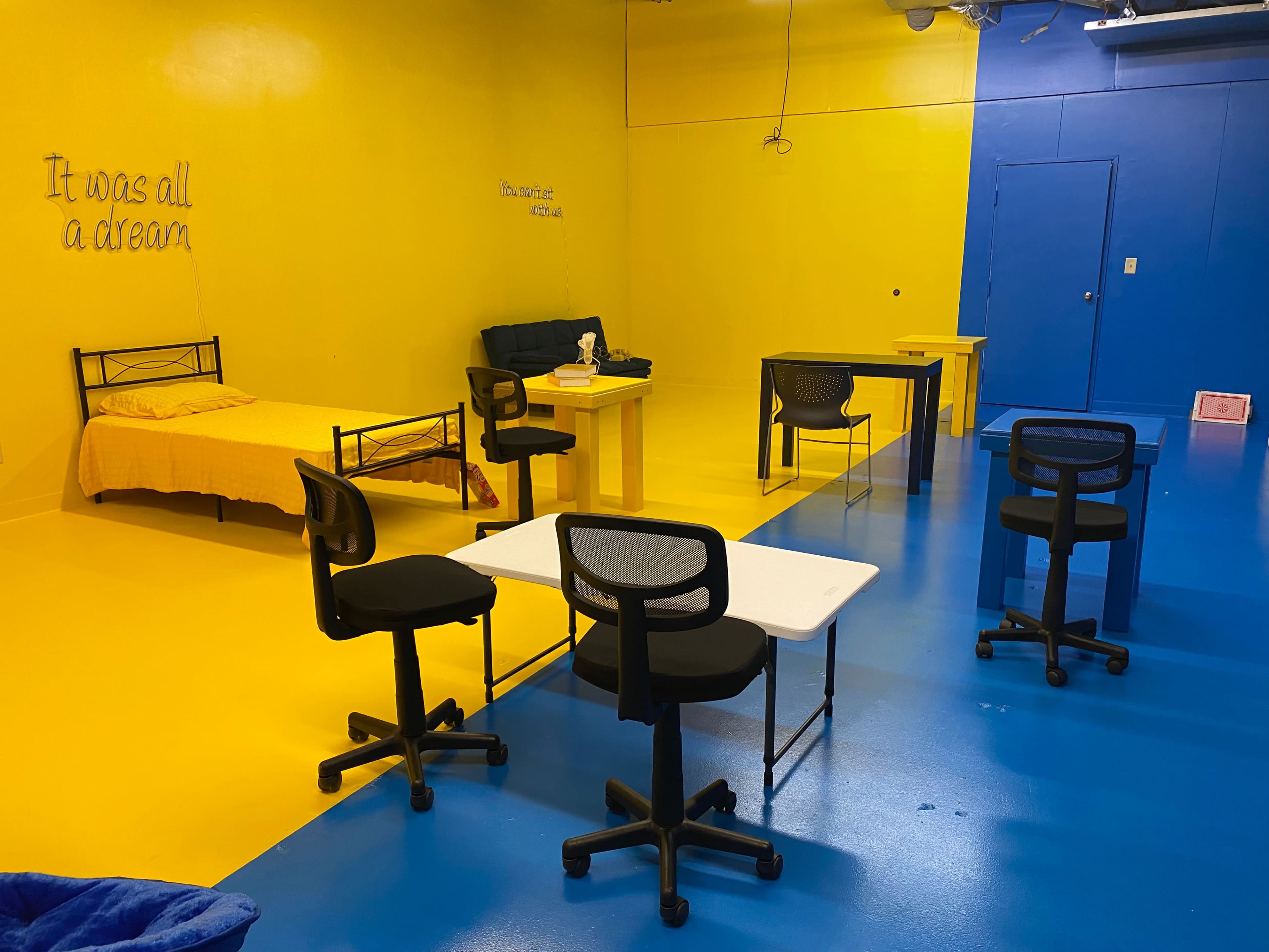 The room features a yellow and blue color scheme with a bed, a couch, several tables, and chairs arranged throughout the space.