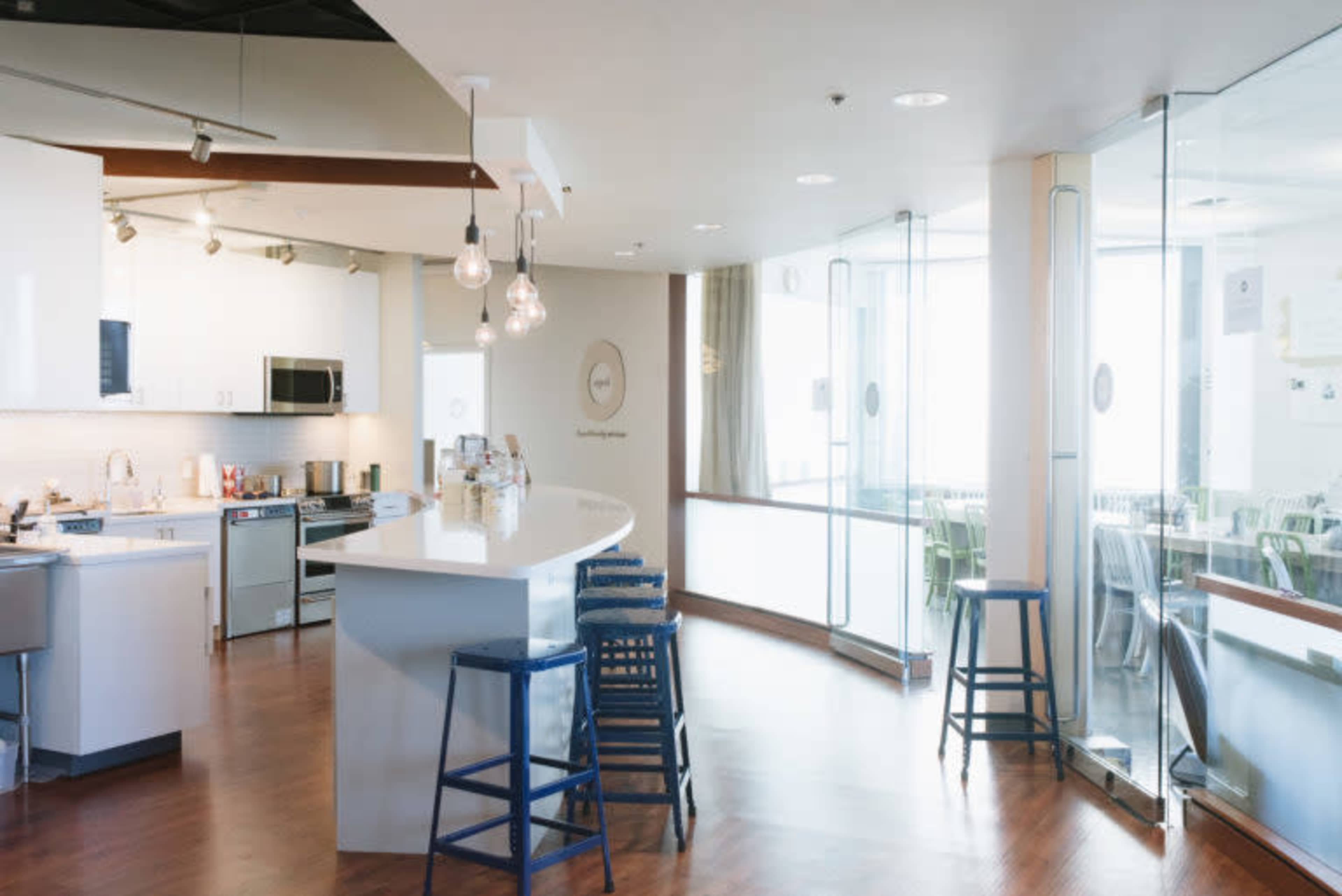 The image shows a modern kitchen area with a curved counter, bar stools, and large windows allowing natural light to enter.