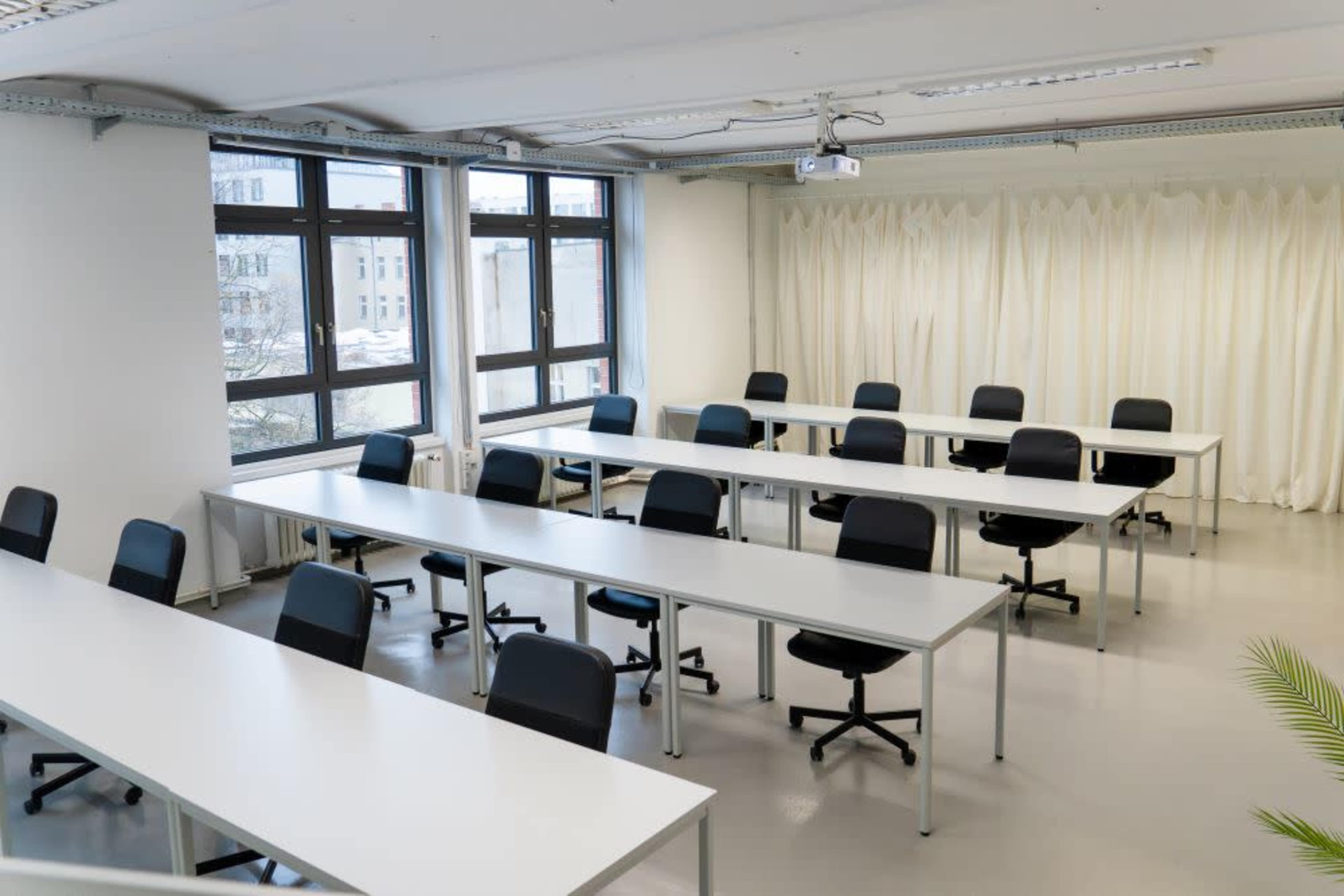 The image shows a classroom with multiple white tables arranged in rows and black chairs, illuminated by natural light from large windows.