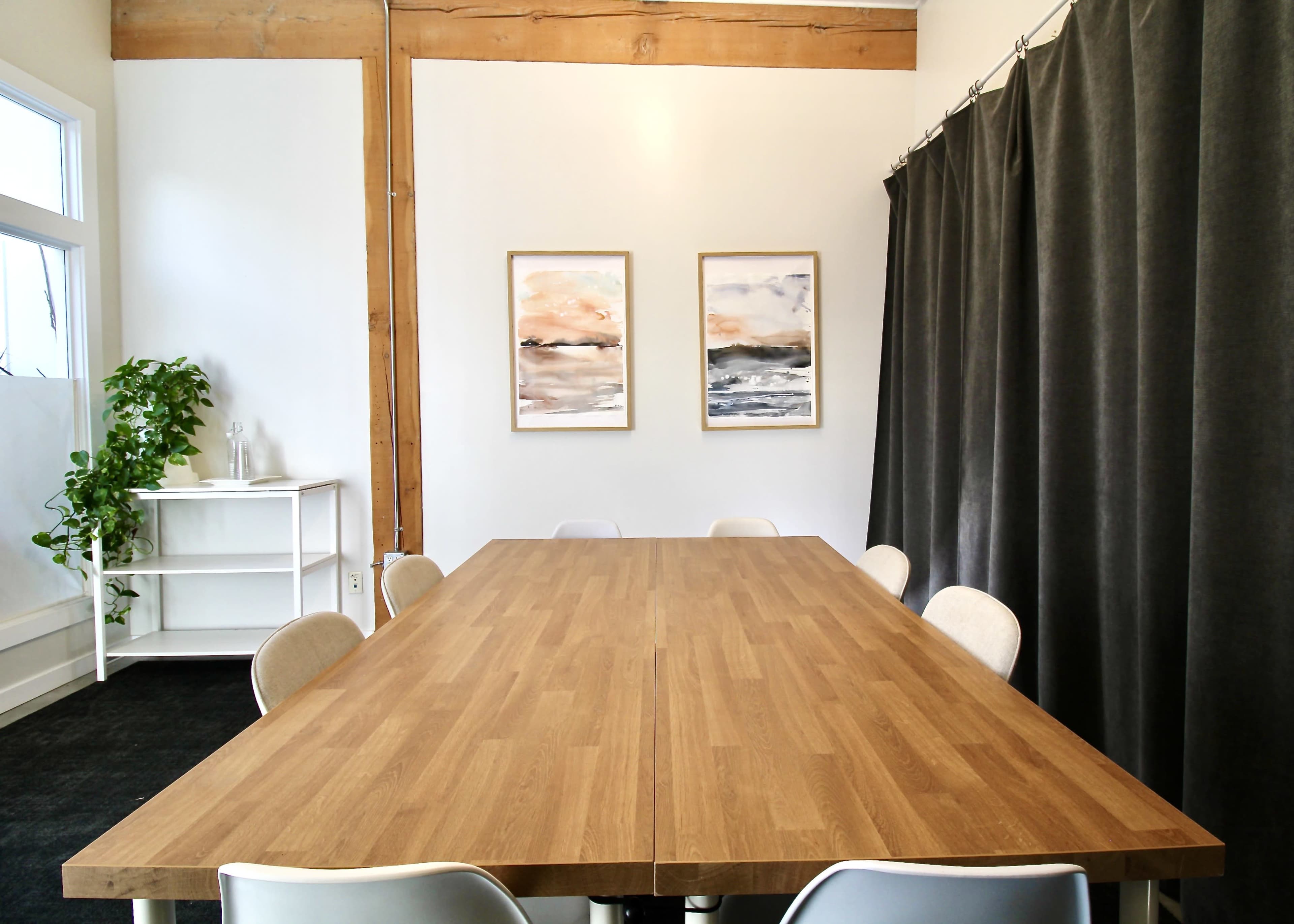 The image shows a large, wooden conference table surrounded by white chairs in a well-lit room with two framed artworks on the wall and a potted plant in the corner.
