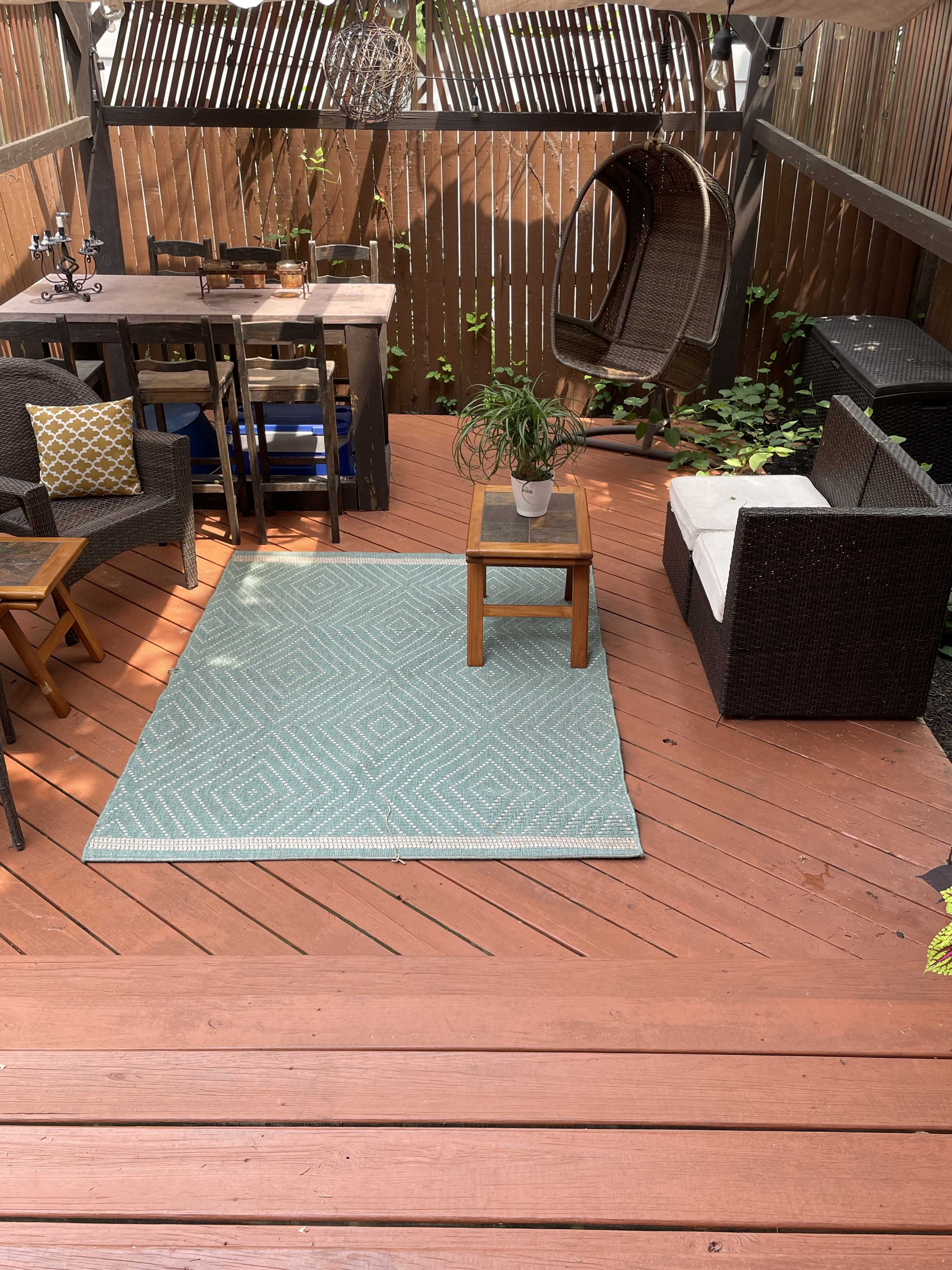 A patio area with wooden decking features a teal geometric rug, a small wooden coffee table with a plant, and outdoor seating arrangements including a hanging chair and a couch.
