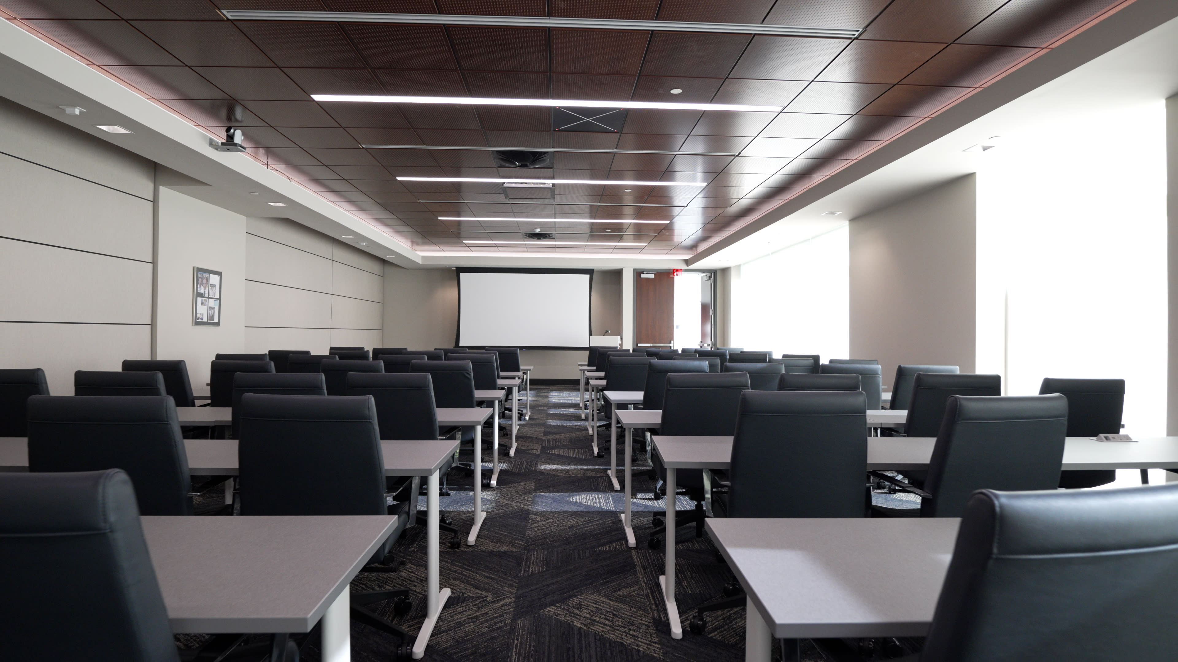 A spacious meeting room is set up with rows of gray tables and black chairs facing a large projection screen.