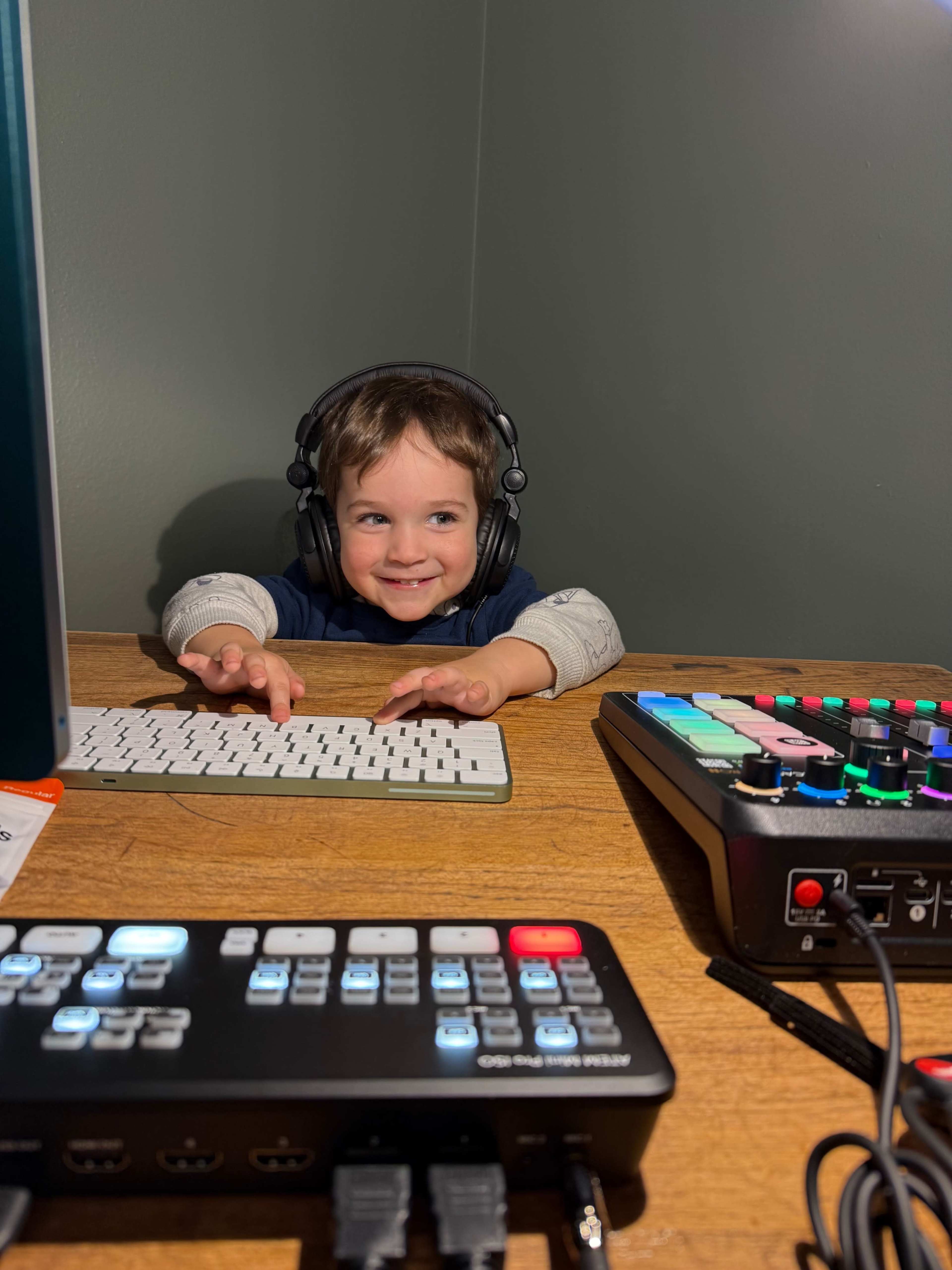 A young child wearing headphones sits at a wooden desk, reaching toward a computer keyboard and an array of electronic music equipment.