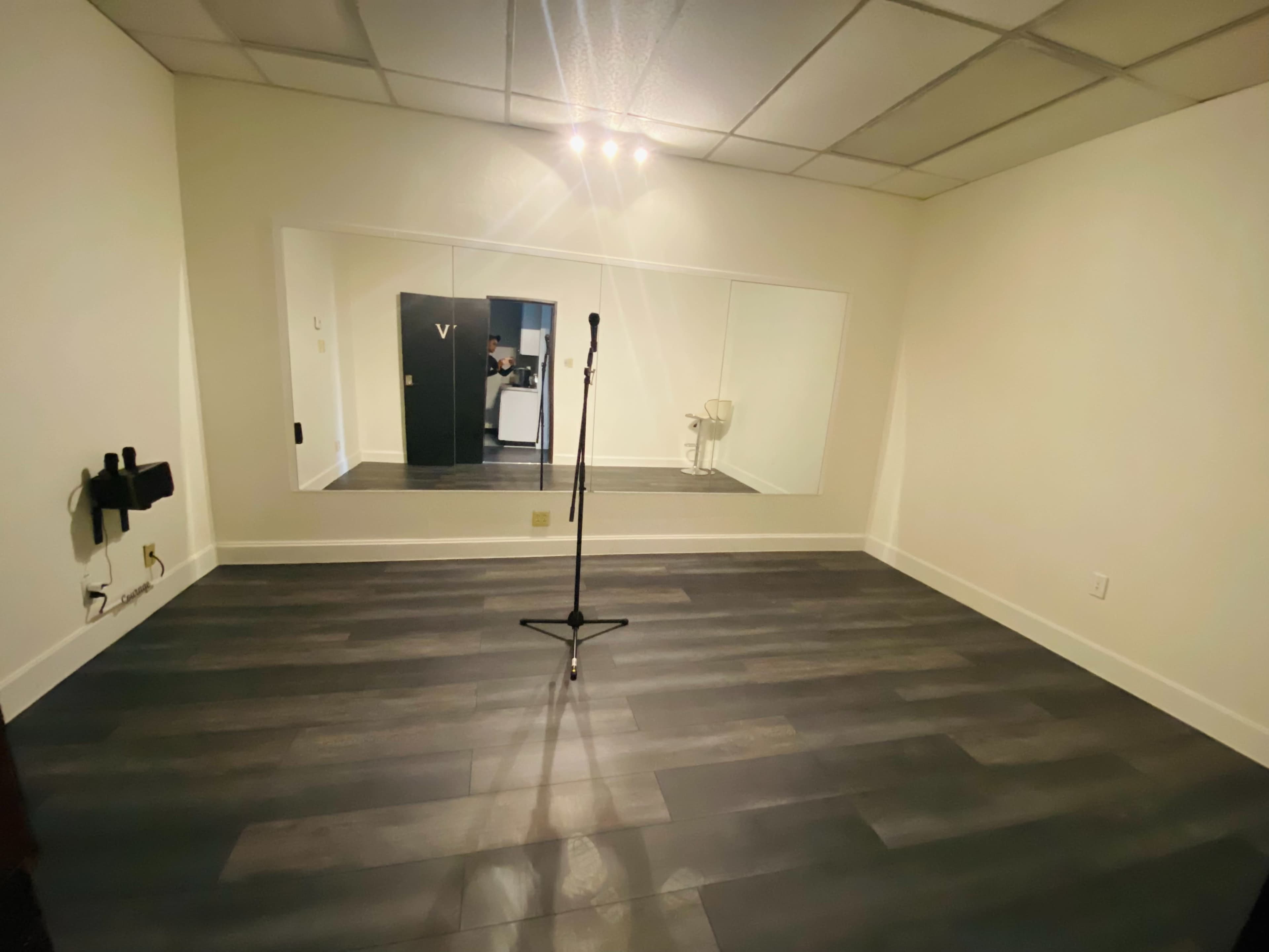 The image shows a small, empty room with a microphone stand in the center and a large mirror on one wall.
