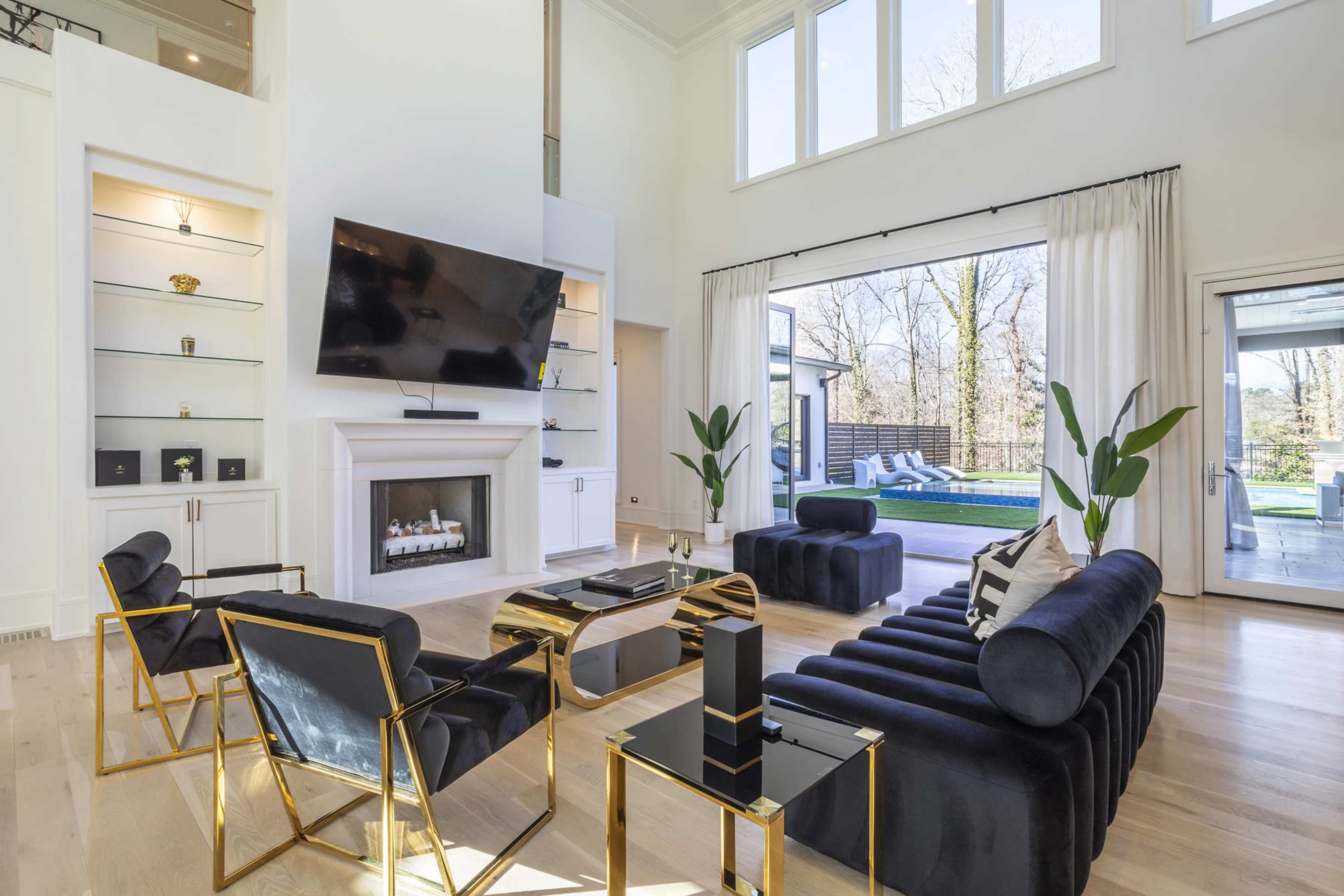 A modern living room with a large television mounted above a white fireplace, featuring black velvet sofas and gold accent chairs, along with large windows that overlook a patio area.