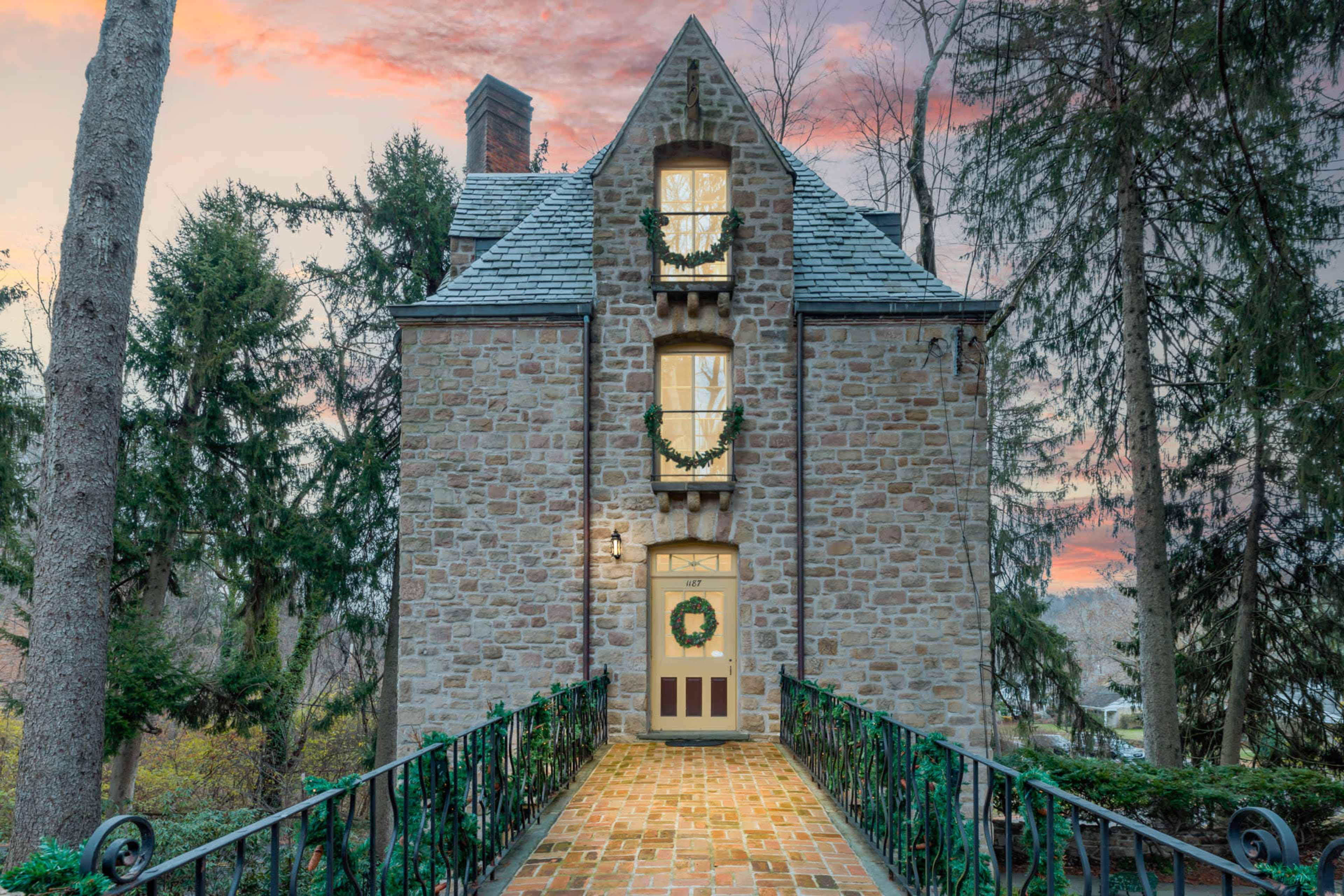 A stone house with a triangular roof is situated at the end of a brick pathway flanked by trees and adorned with seasonal decorations.