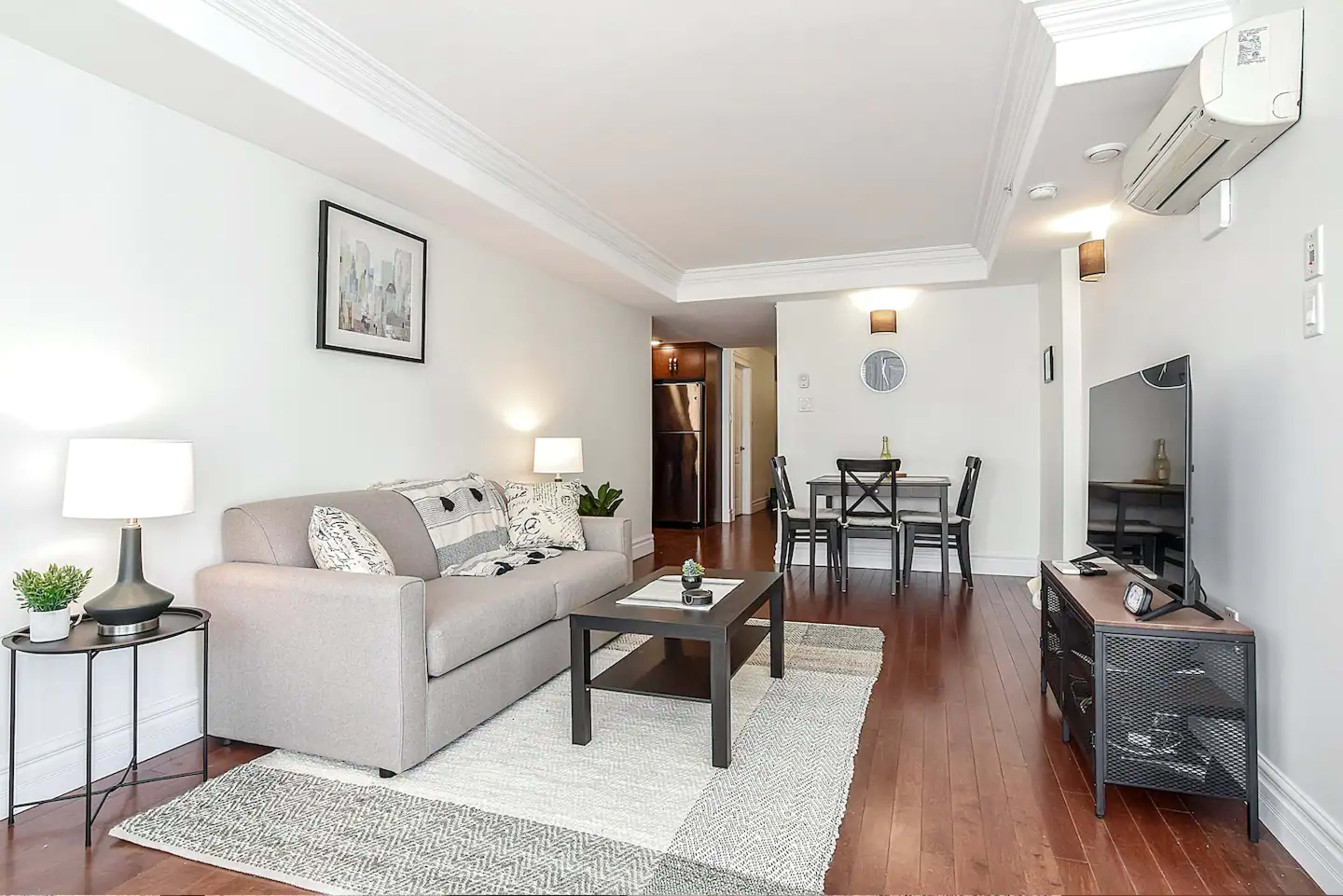 A living area features a couch, coffee table, and television, with a dining space and kitchen visible in the background.