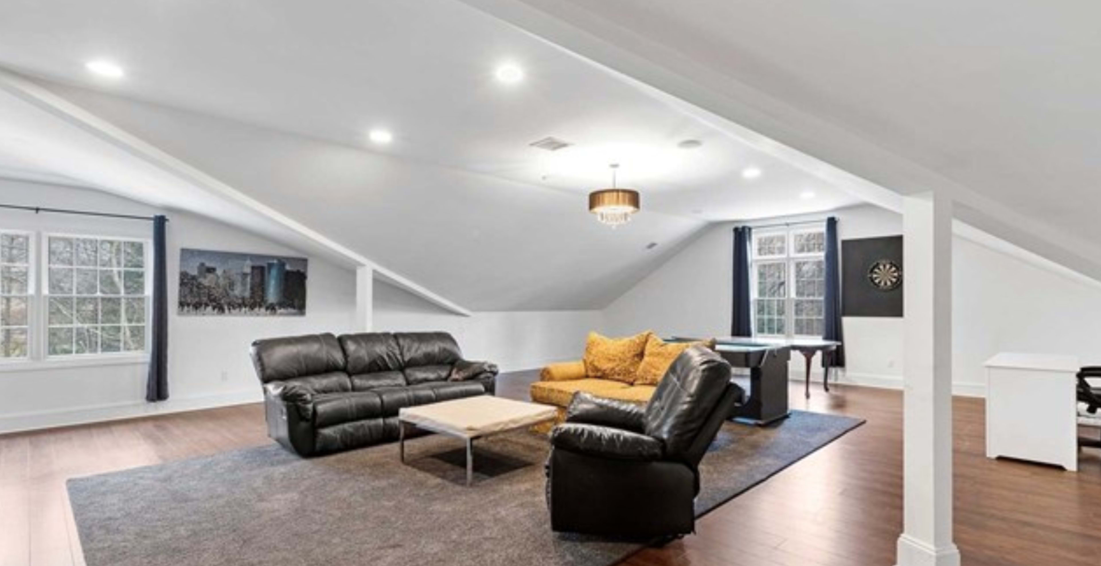 A spacious attic living area with two leather sofas, a coffee table, a bright chandelier, and large windows providing natural light.
