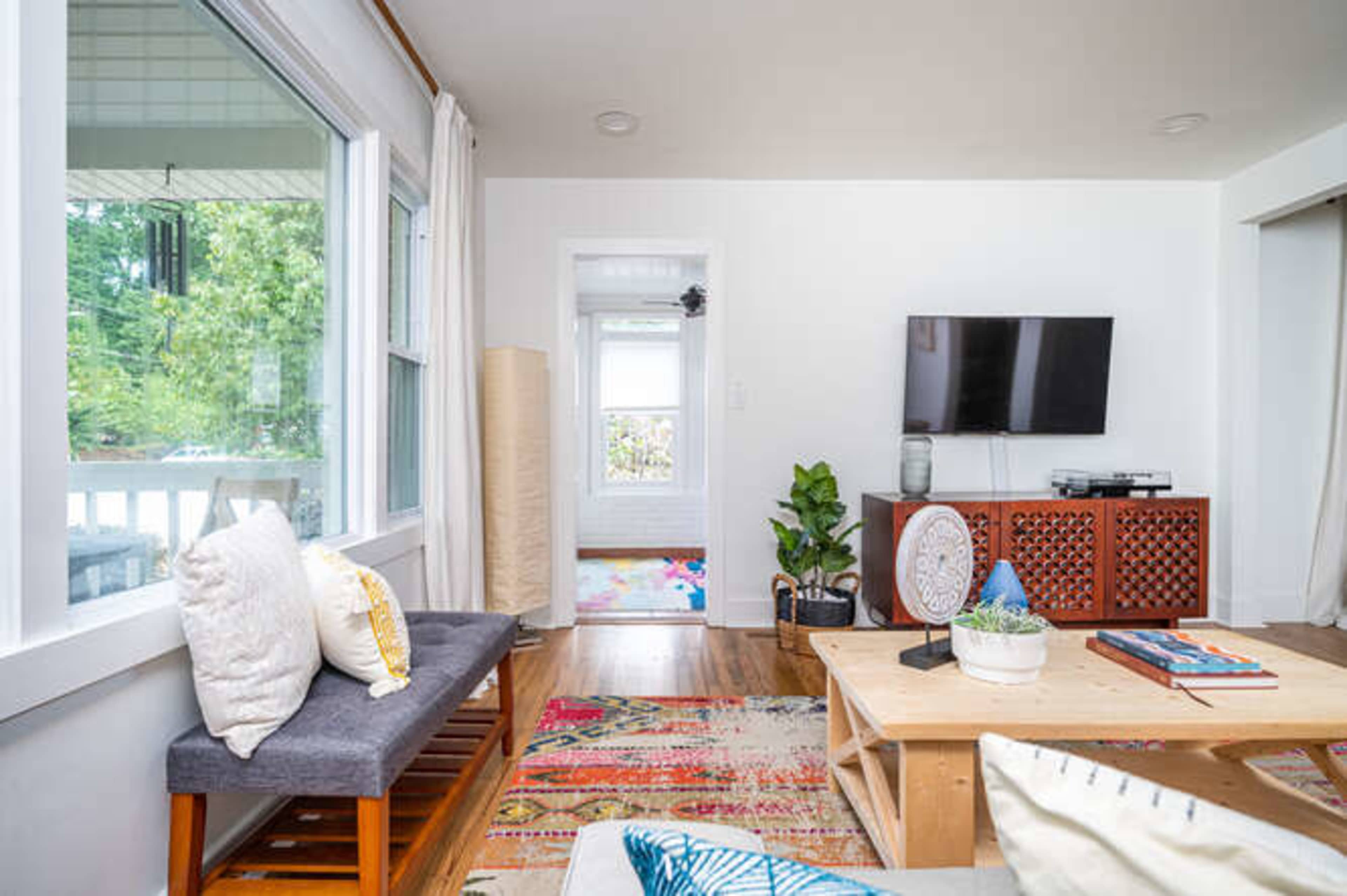 The image shows a bright living room with a television mounted on the wall, a coffee table, and a seating area, featuring large windows that allow natural light to enter.