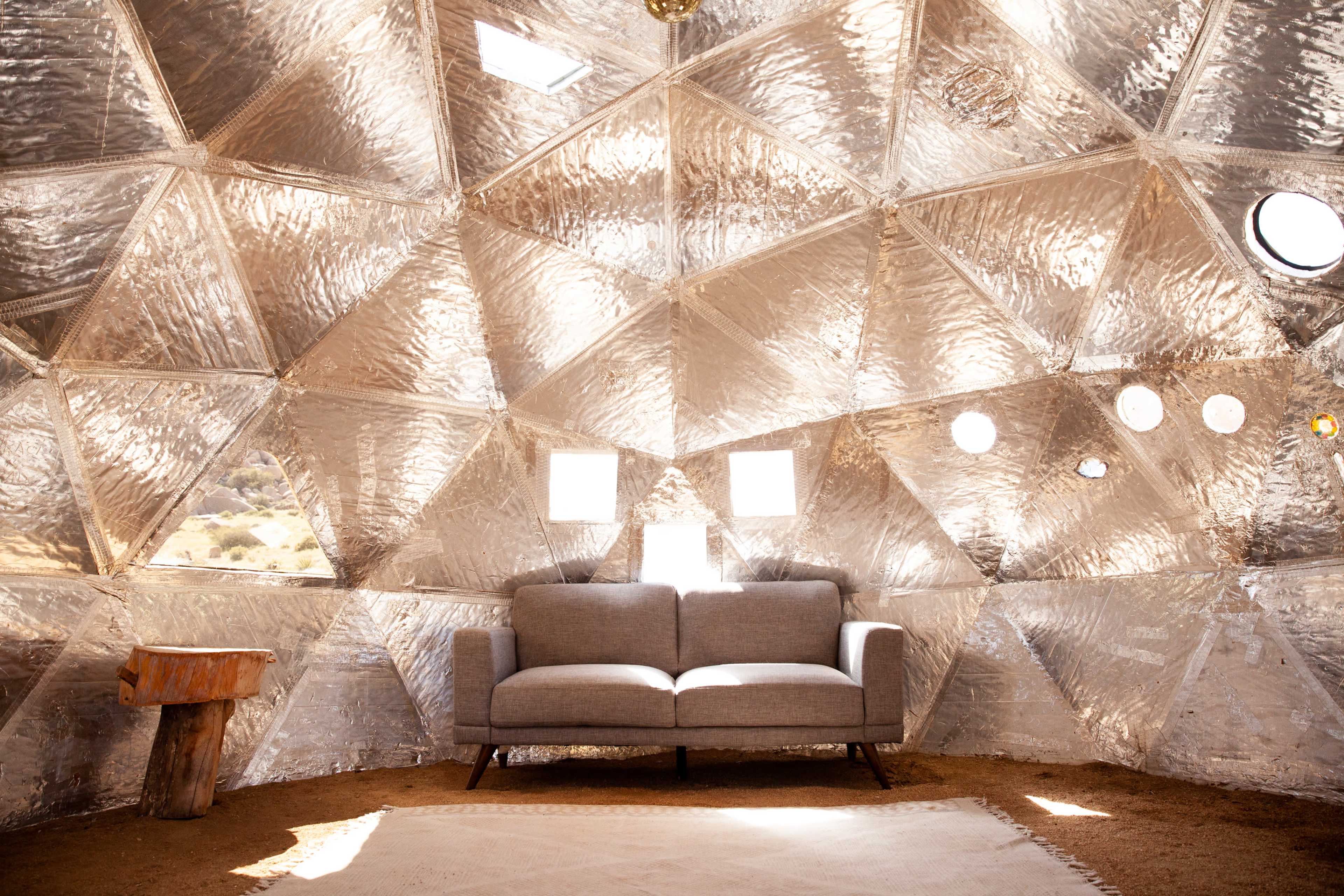 The interior of a geodesic dome features a gray sofa on a textured floor, surrounded by reflective walls and circular windows.