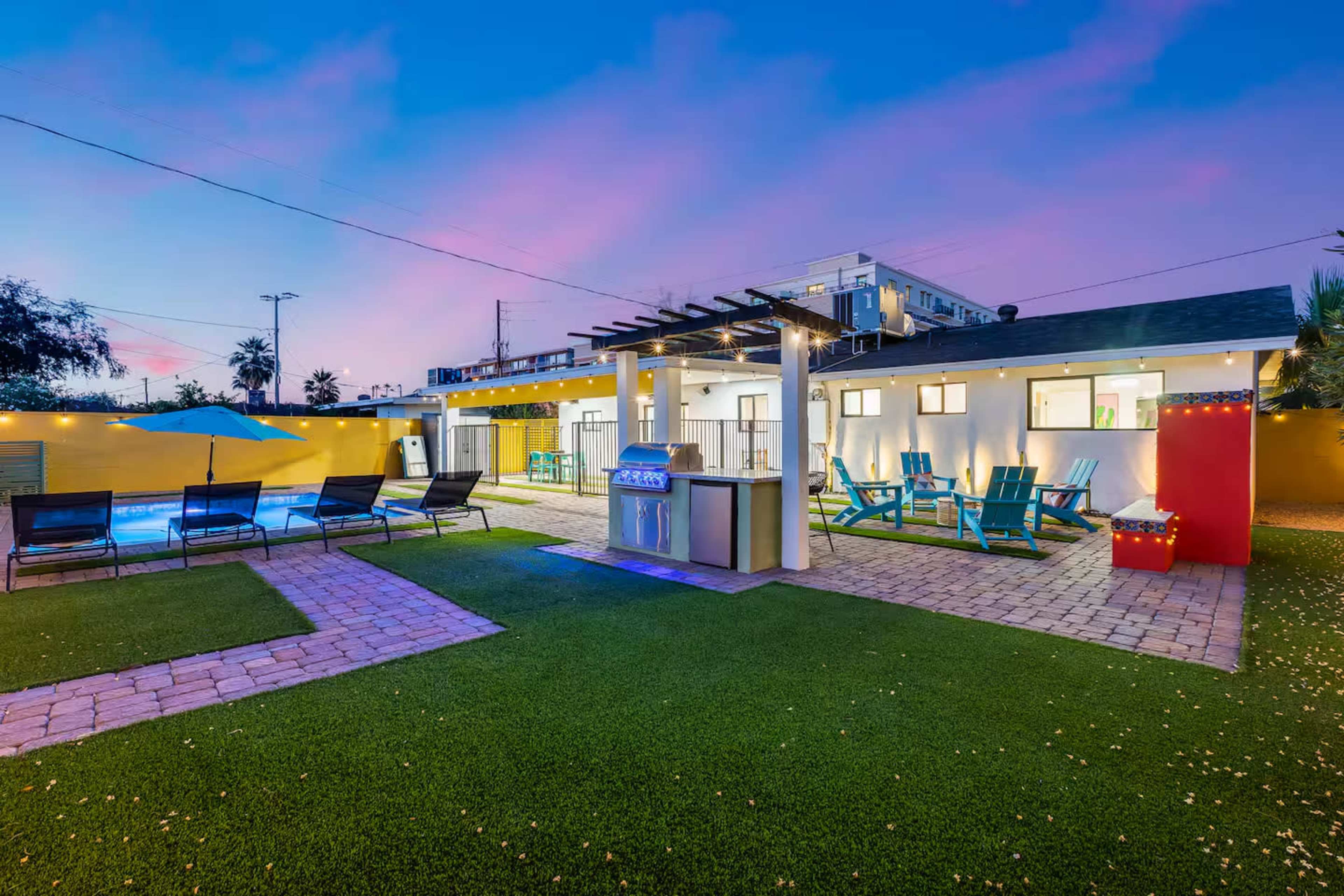 The image depicts a backyard area featuring a swimming pool, lounge chairs, colorful seating arrangements, and a barbecue area, with buildings in the background under a twilight sky.