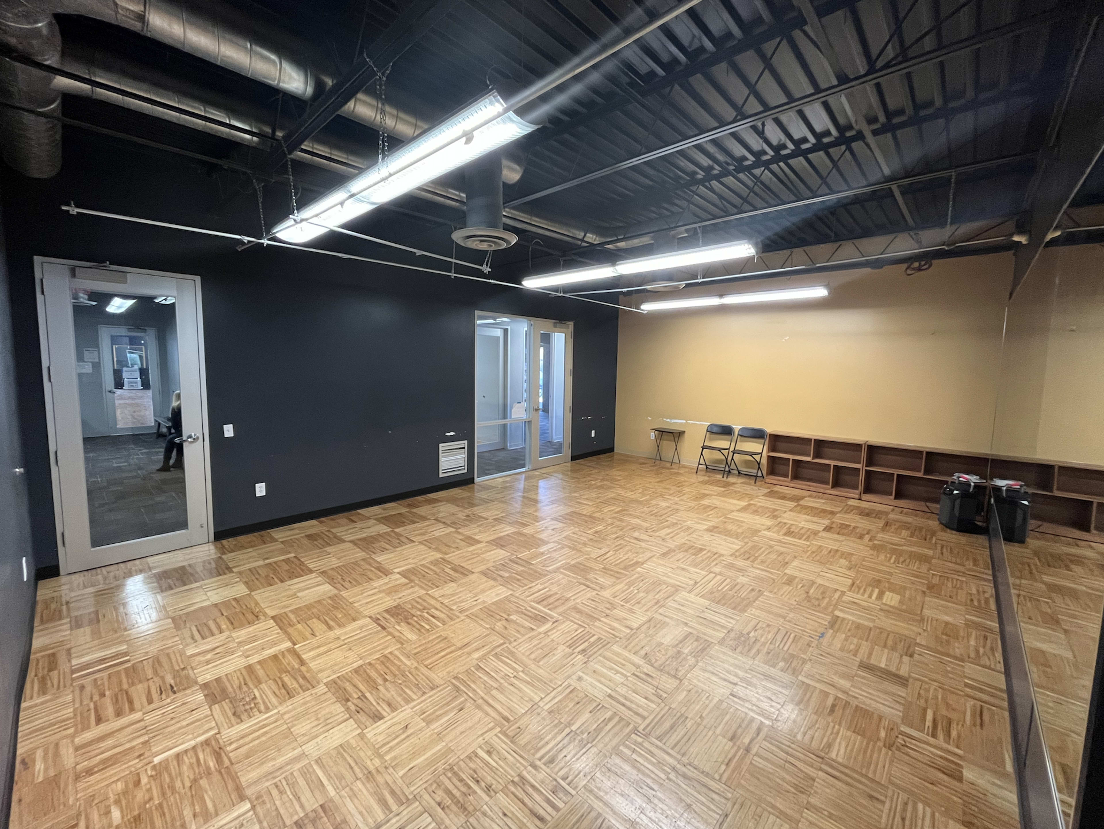 The image shows an empty room with wood-paneled flooring, black walls, and a small adjoining space with a glass door.