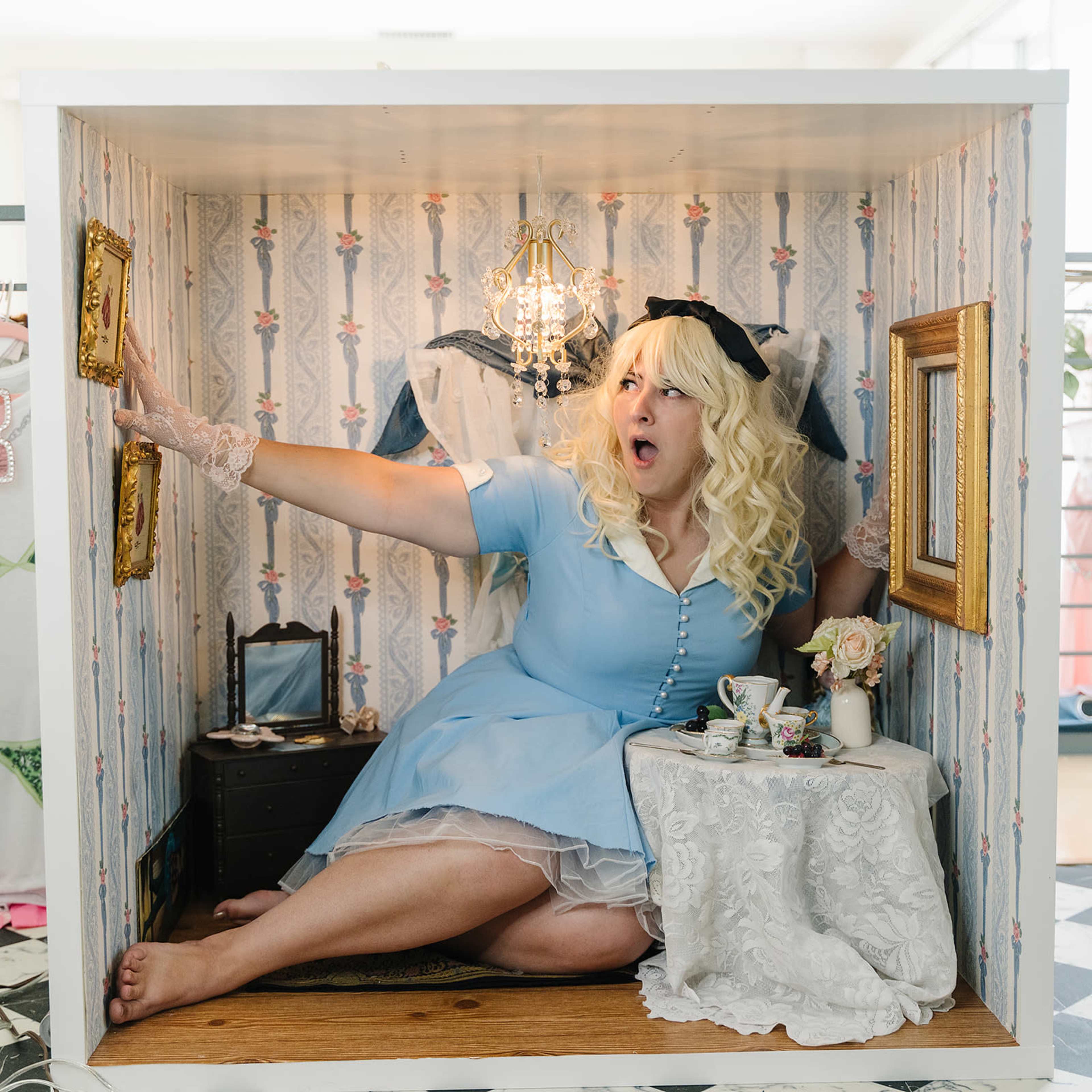 A woman in a blue dress sits in a small box-like setting, decorated with floral wallpaper, holding a surprised expression while surrounded by vintage items.