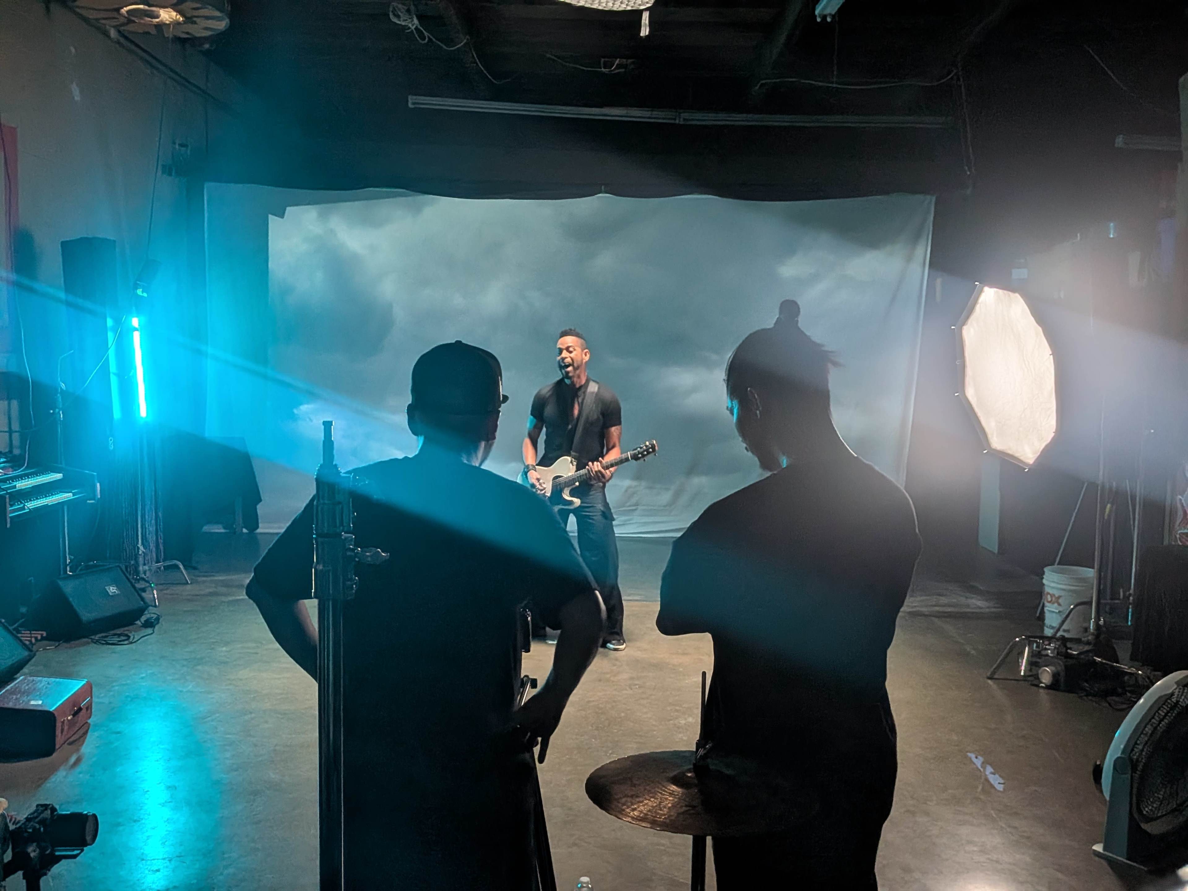 A musician plays guitar on a stage with a cloudy backdrop while two crew members observe from behind.