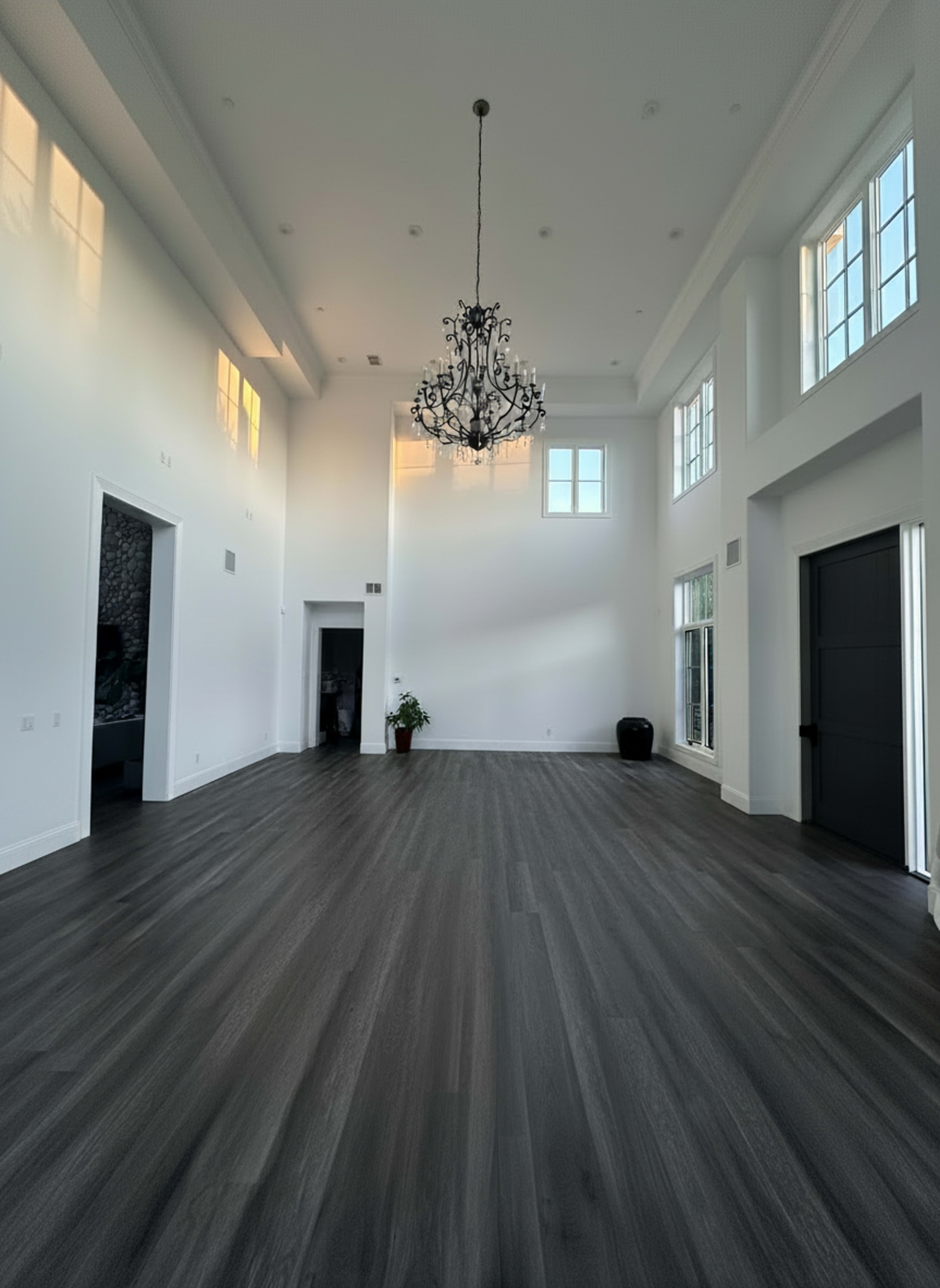 A large, brightly lit room features high ceilings, large windows, a chandelier, and dark wooden flooring.