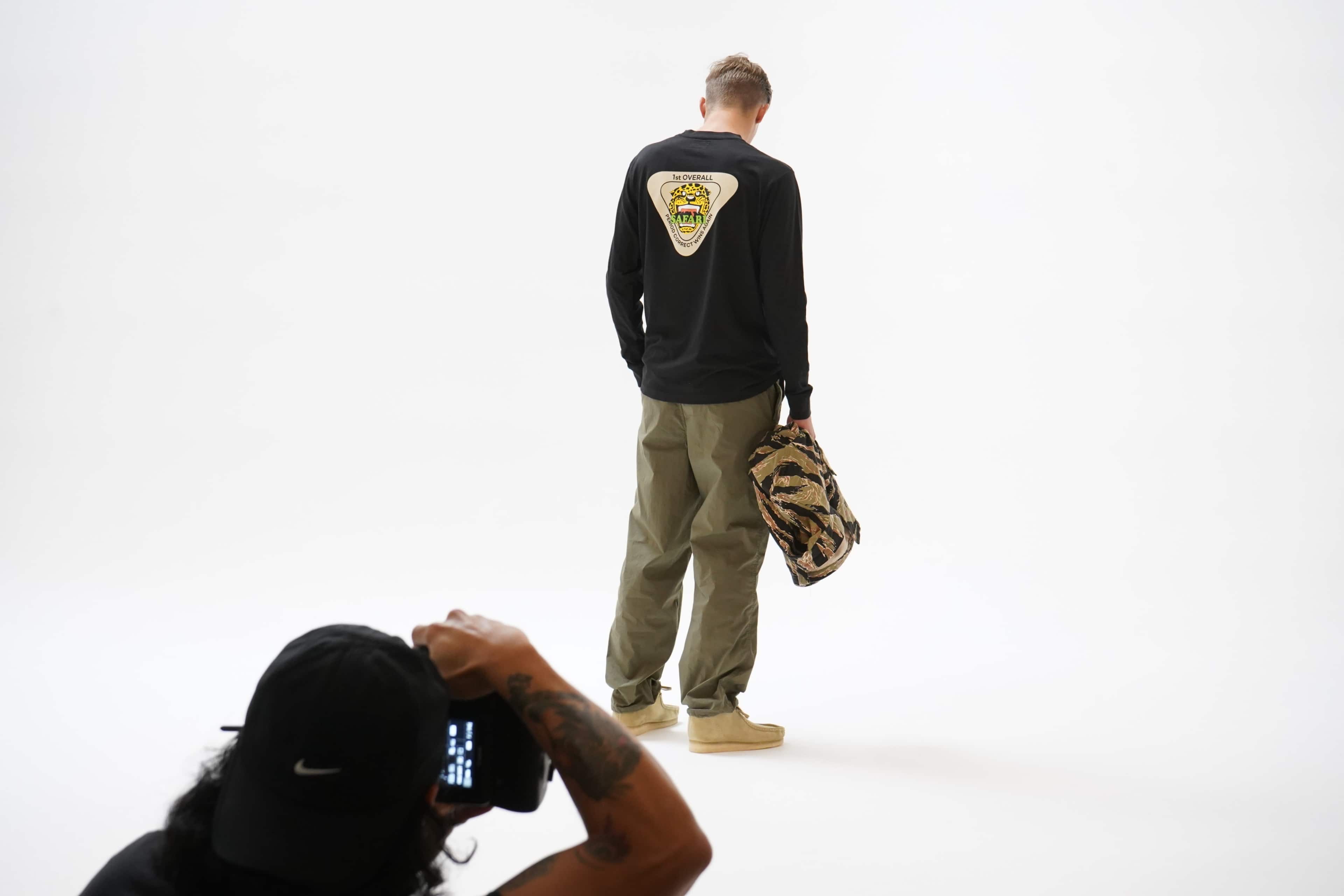 A person wearing a black long-sleeve shirt and olive green pants stands with their back to the camera while holding a camouflaged bag, as another individual takes a photograph.