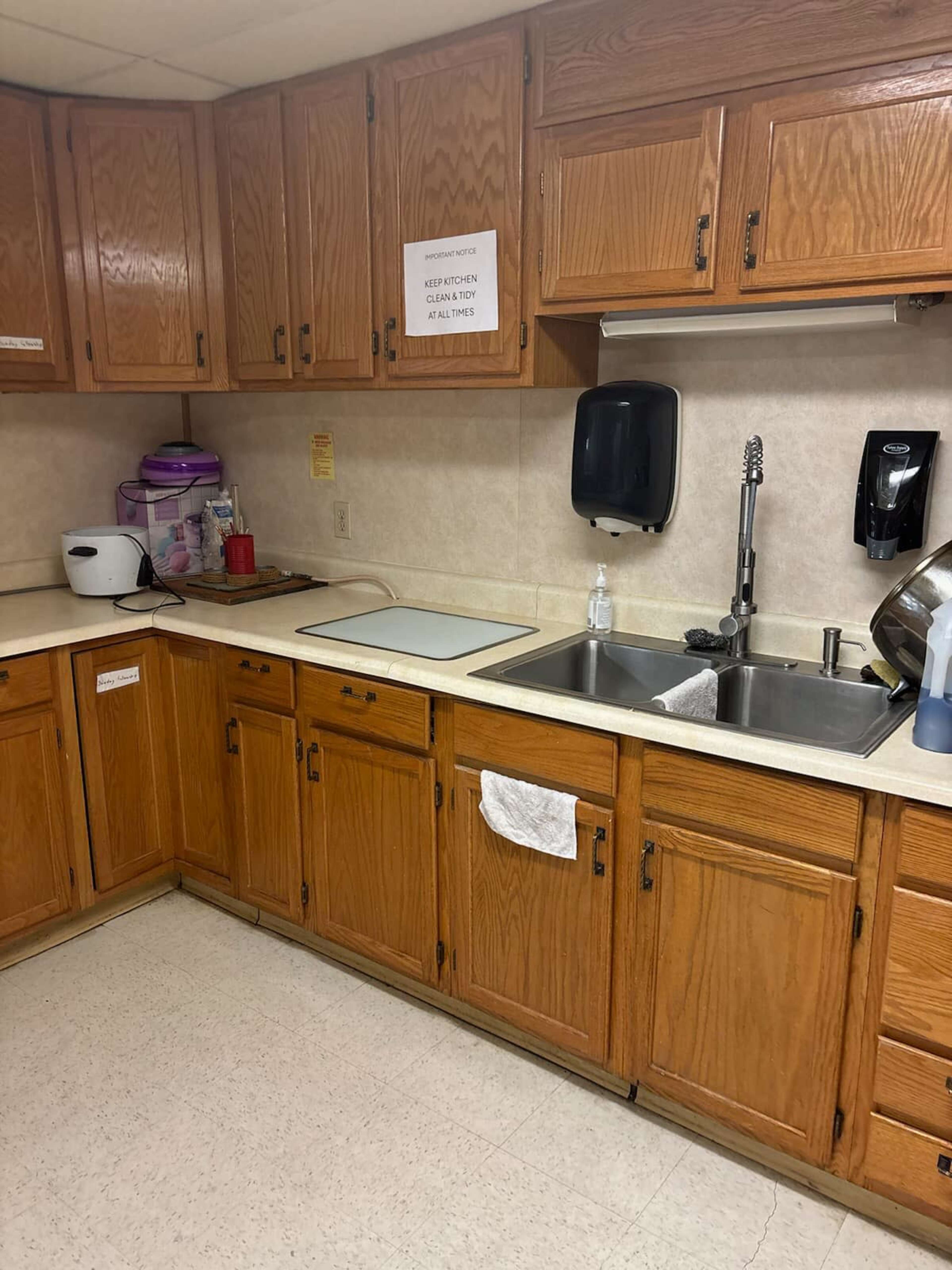 The image shows a kitchen with wooden cabinetry, a sink, a countertop, and various appliances and supplies.