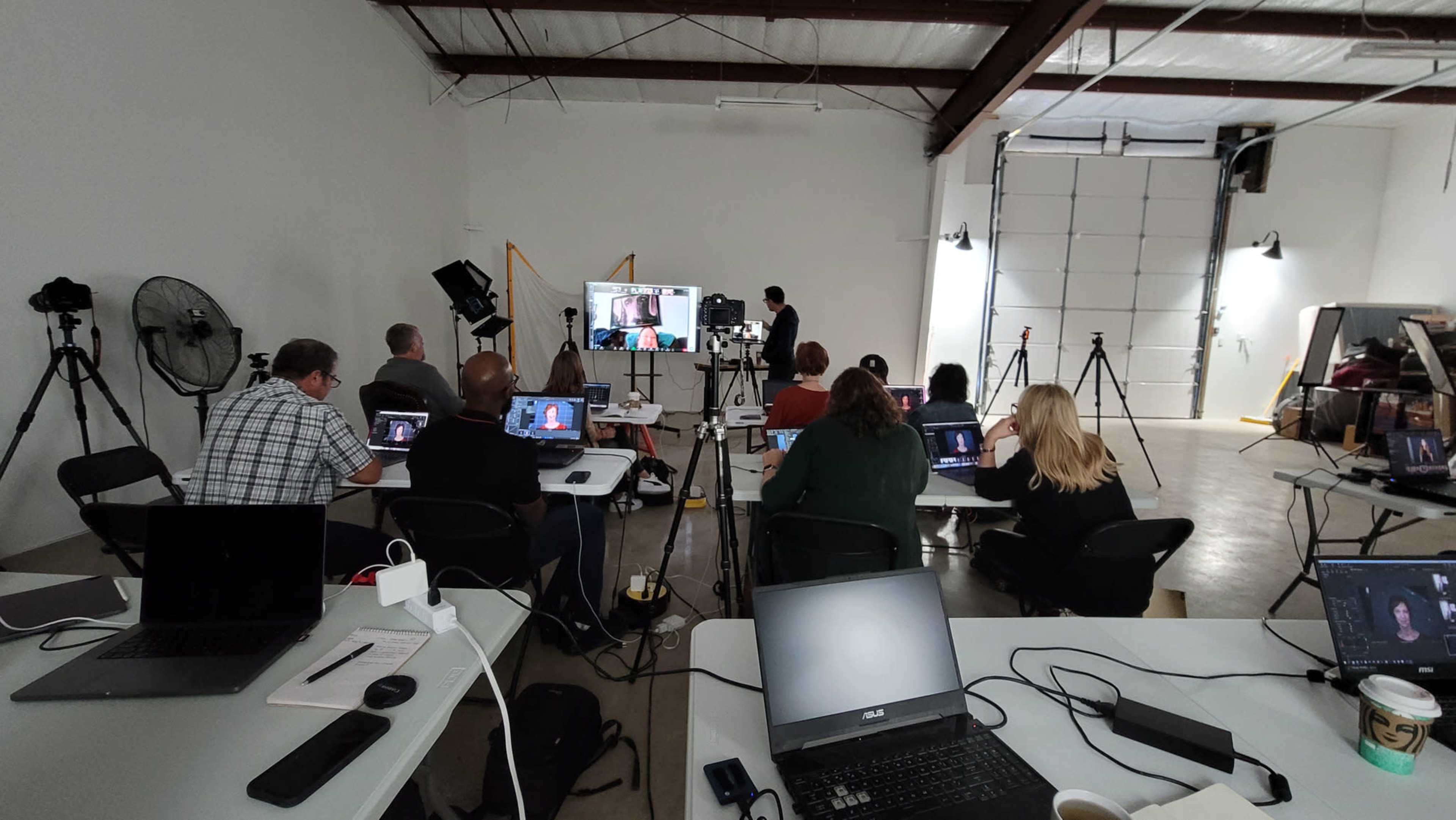 A group of people is seated at tables in a studio, participating in a video conference session with multiple screens and cameras set up around the room.