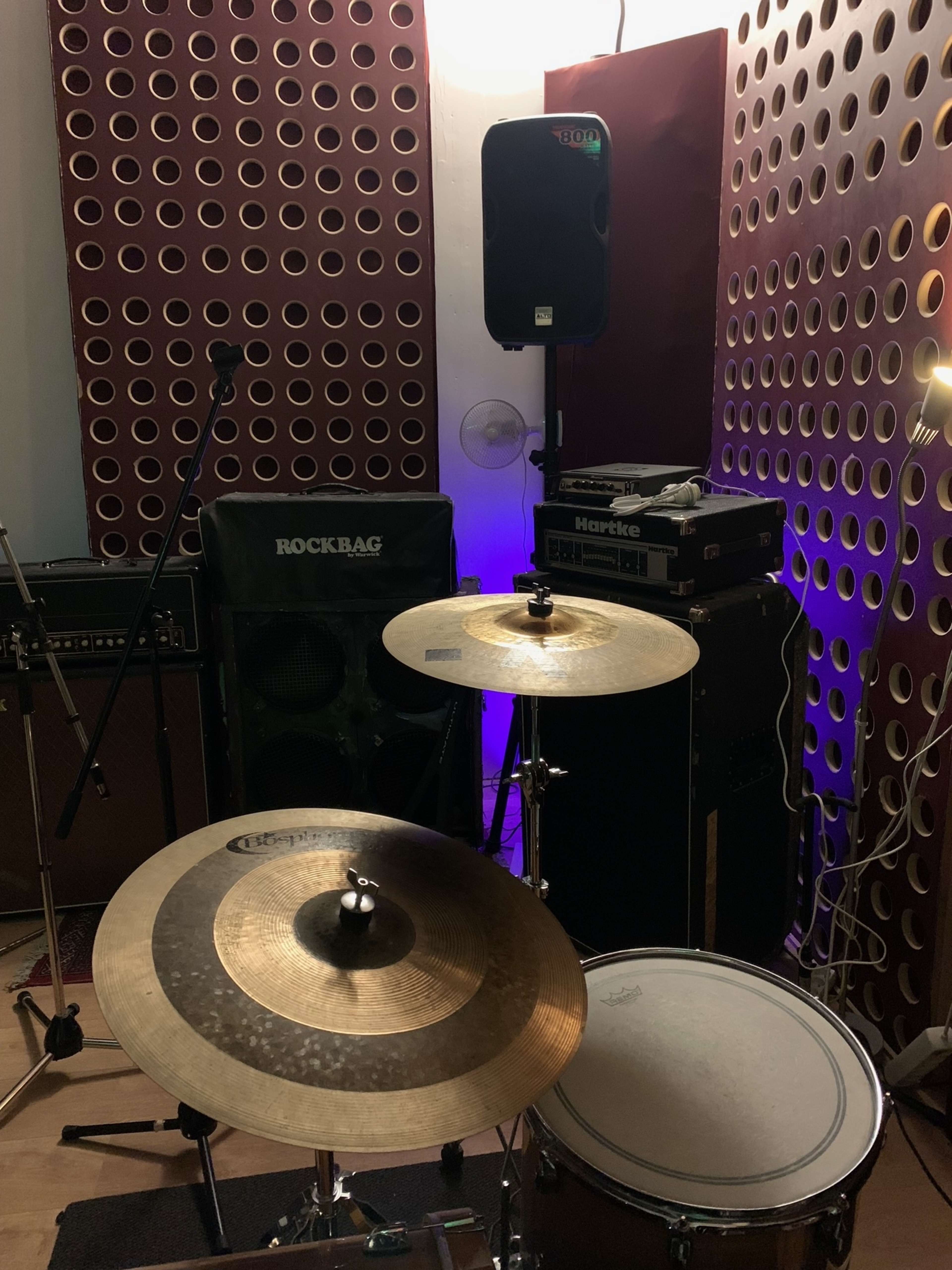 A music rehearsal space with a drum set, amplifiers, a microphone stand, and soundproofing panels on the walls.