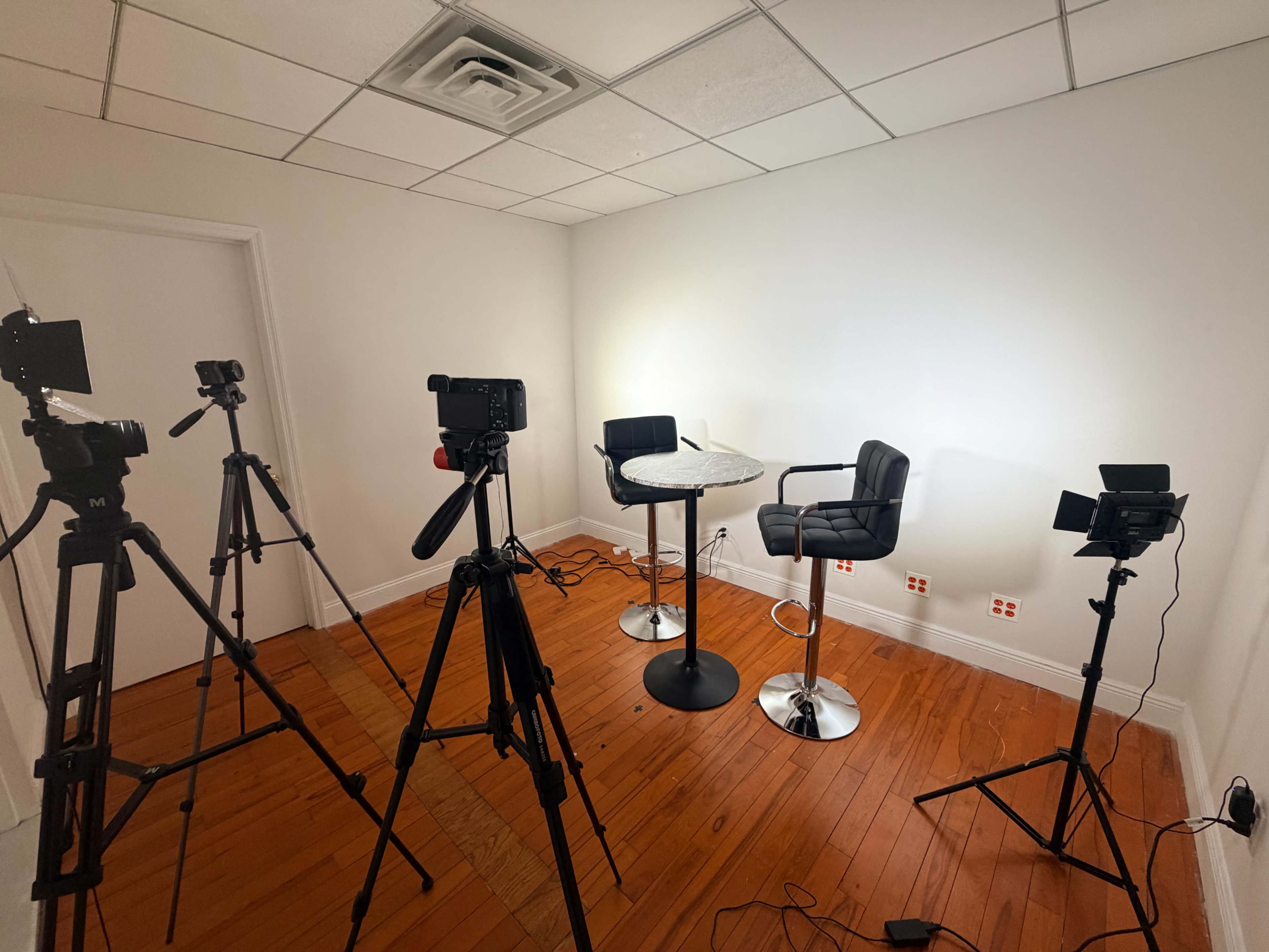 The image shows a small studio setup with two empty barstools, a round table, and multiple cameras on tripods arranged around the space, all against a plain white wall.