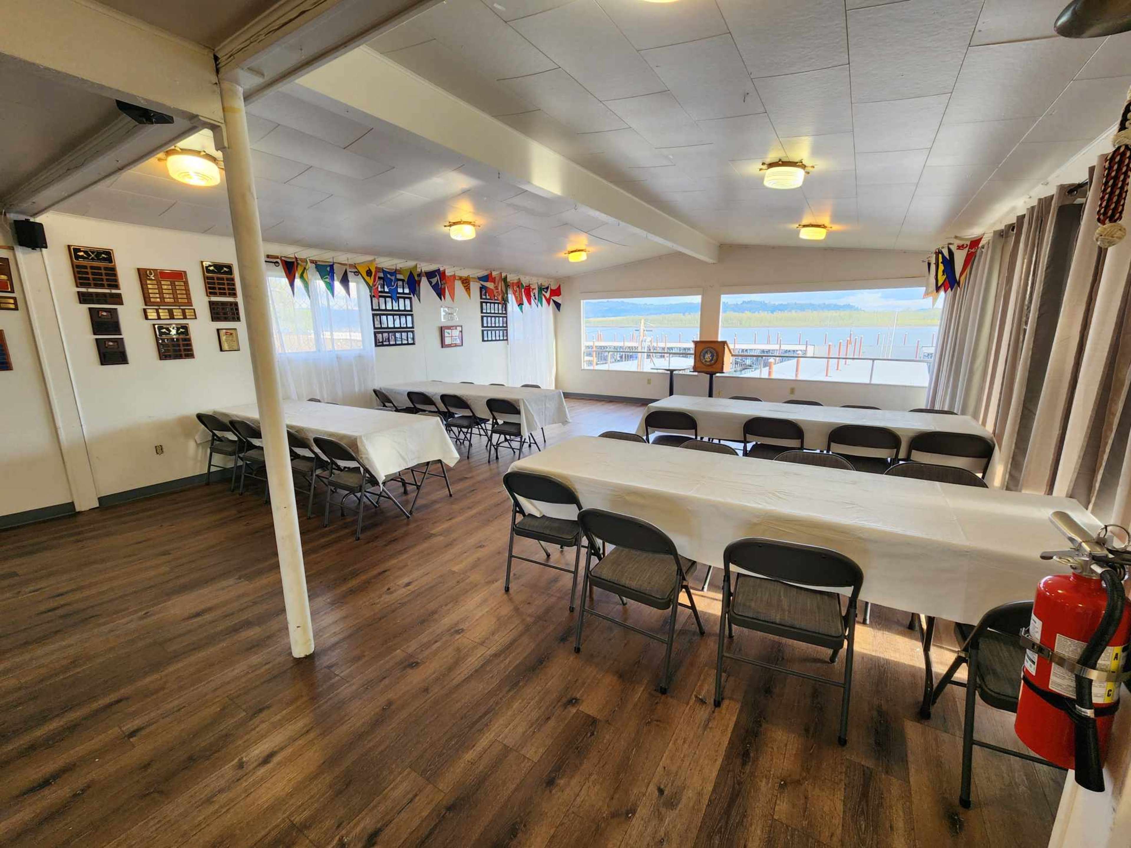 A spacious indoor area with several tables covered in white tablecloths, arranged in a setting with nautical decorations and large windows overlooking a body of water.