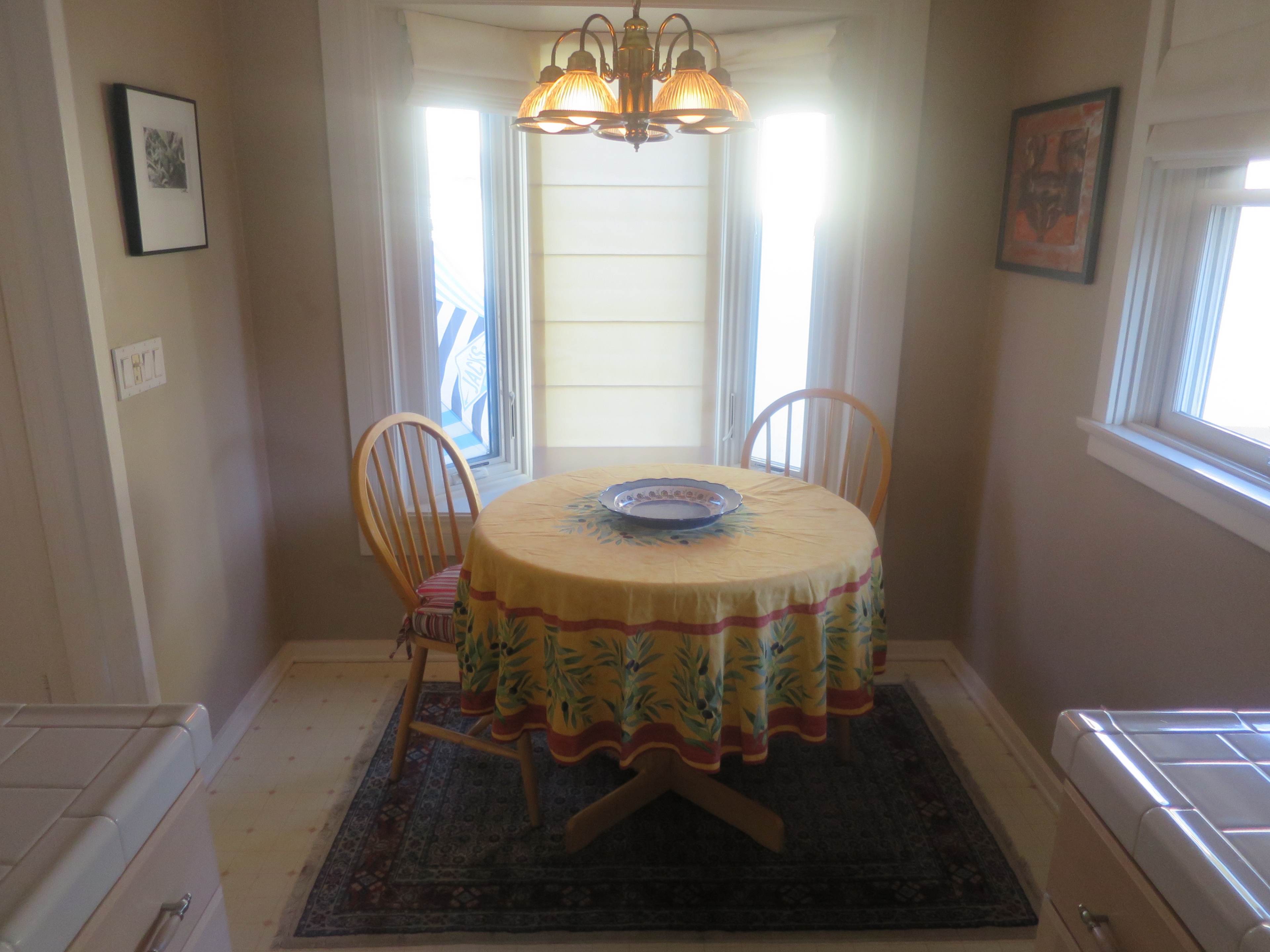 A small dining area features a round table with a yellow patterned tablecloth and two wooden chairs, surrounded by windows allowing natural light.