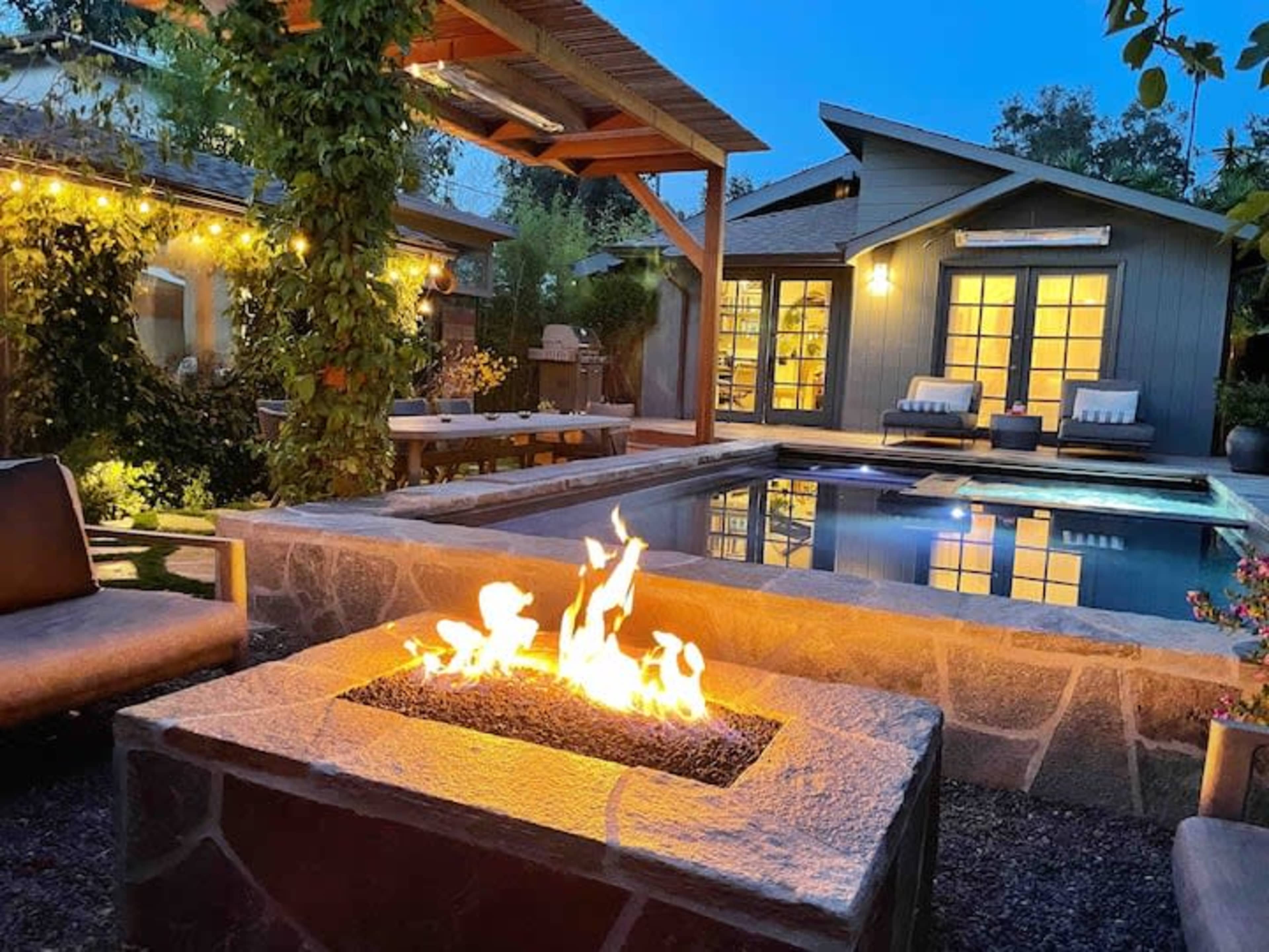 A stone fire pit with flames is positioned near a swimming pool and a modern house, surrounded by patio seating and greenery in the evening light.