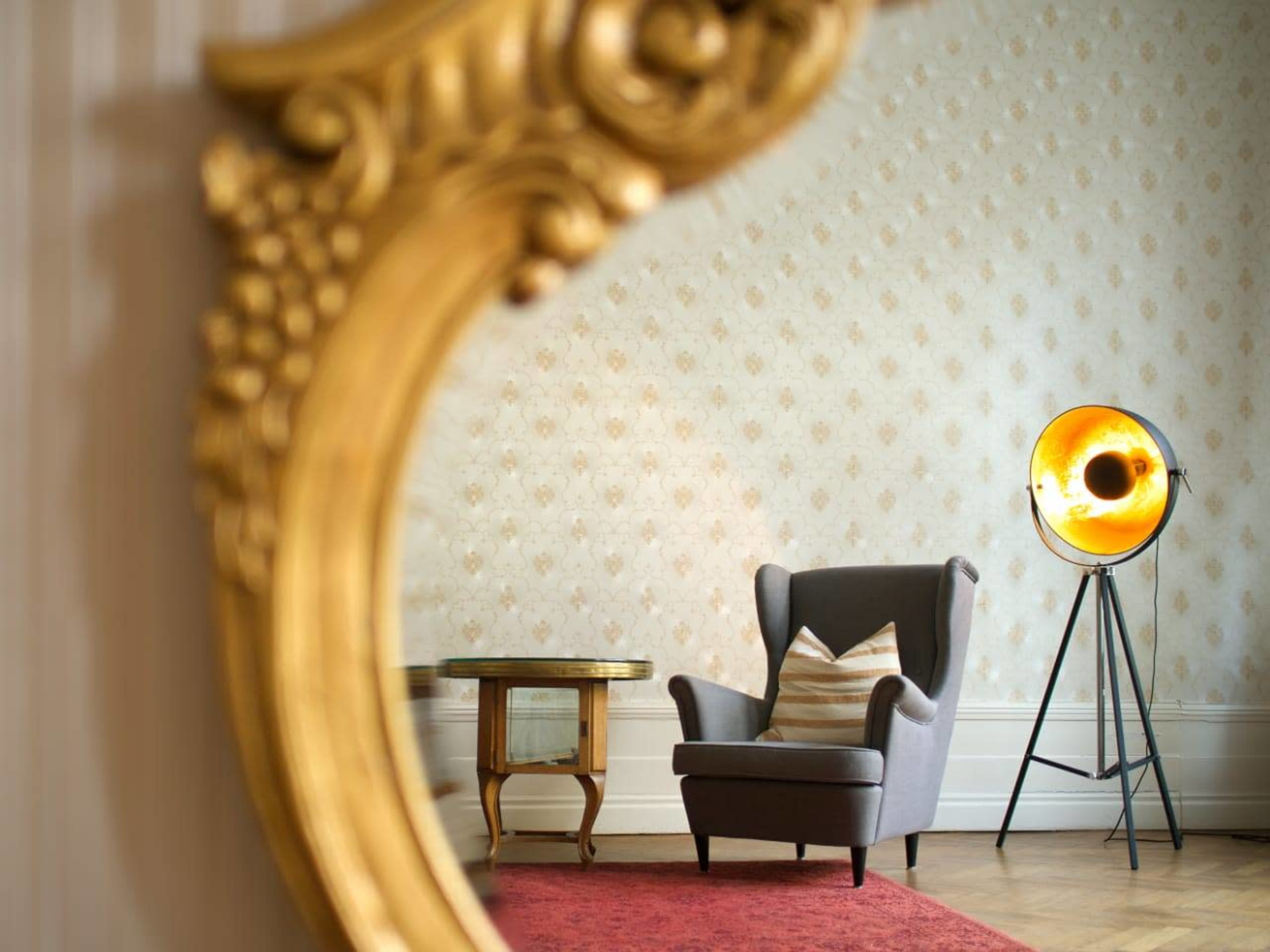 A gray armchair with a striped cushion is positioned next to a modern floor lamp, reflected in a decorative gold-framed mirror against a patterned wallpaper backdrop.
