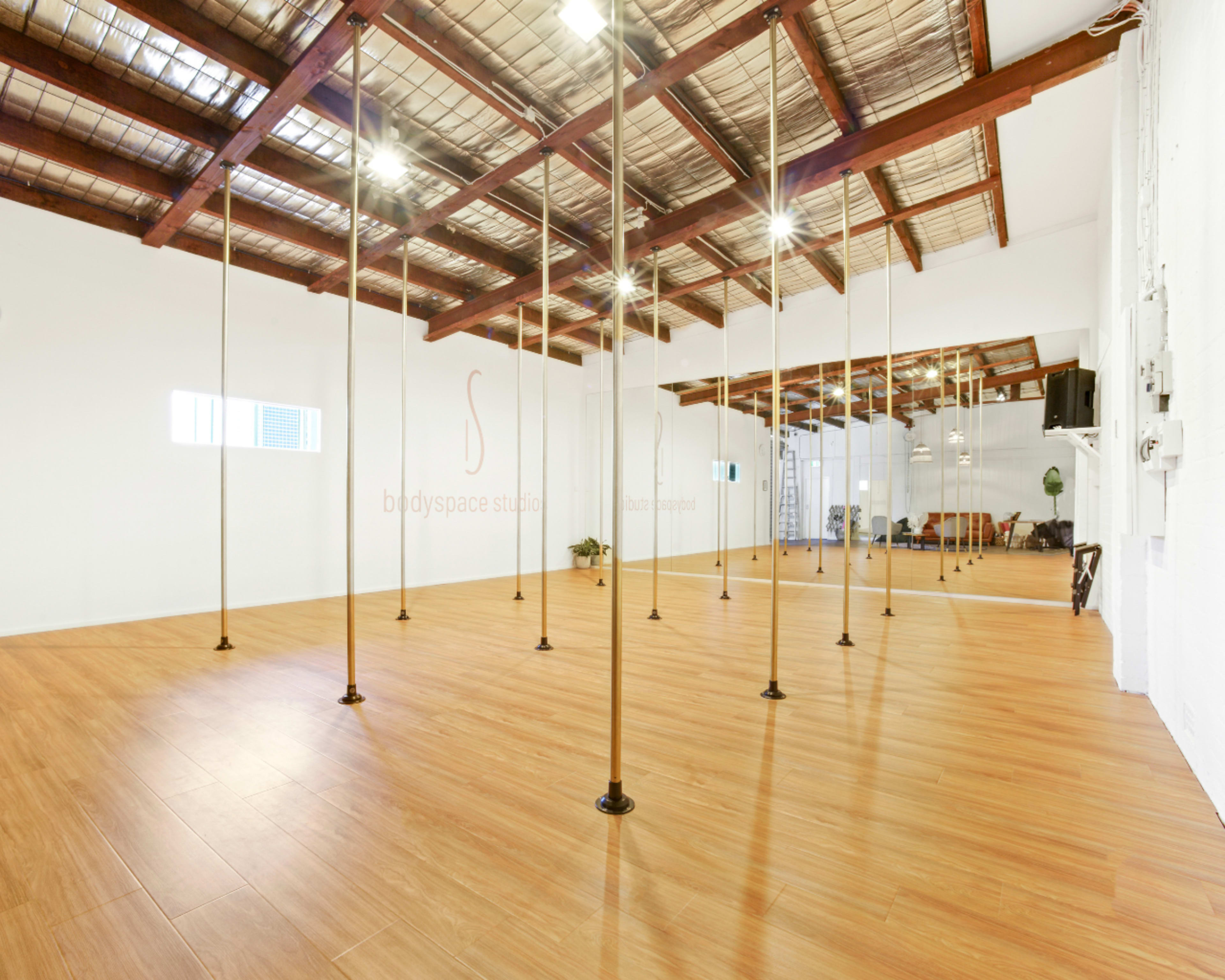 The image shows a spacious studio with wooden flooring, tall poles evenly spaced throughout, and a minimalist design featuring white walls and exposed wooden beams.