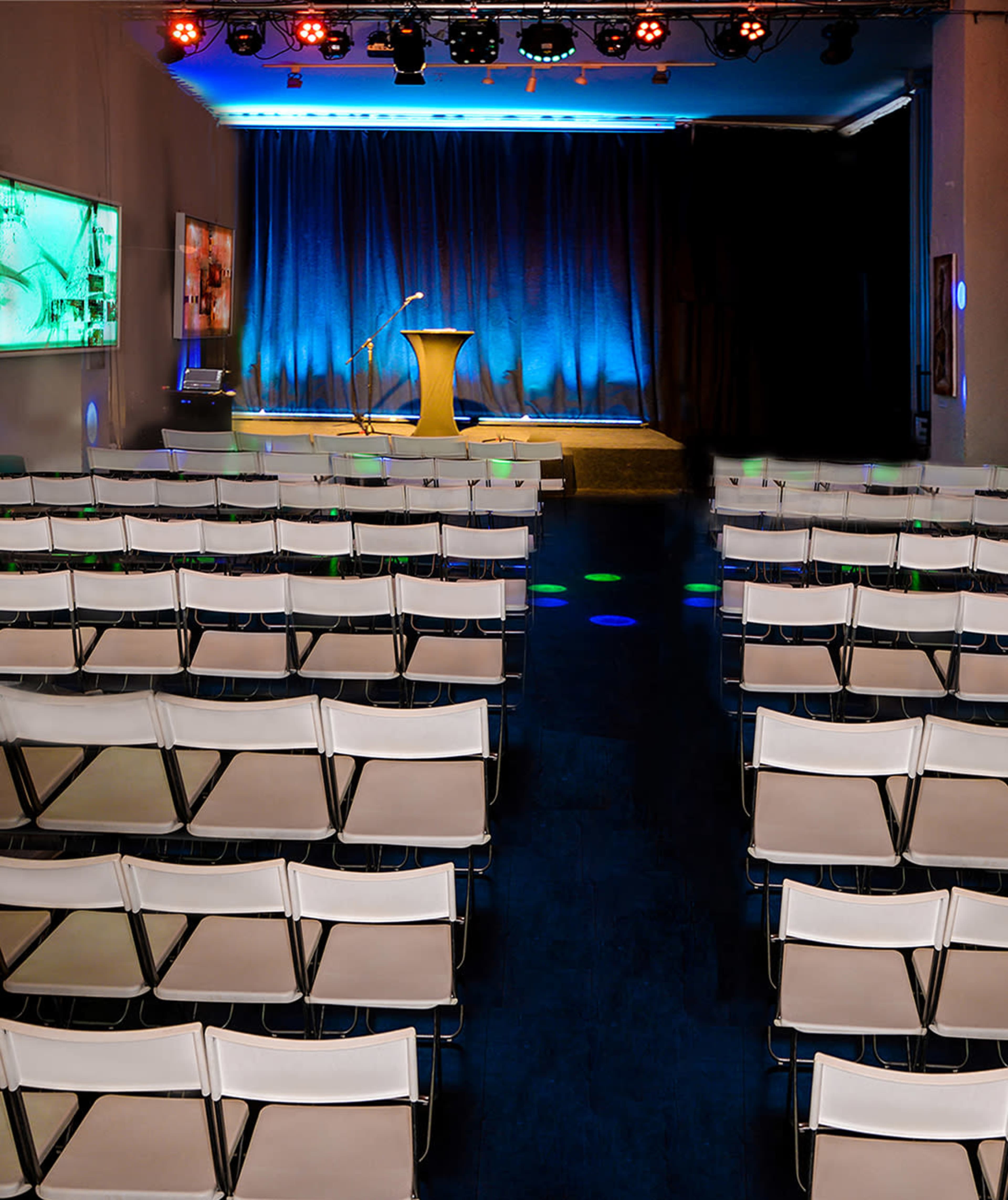 A stage with a podium and microphone is set at the front of a room filled with rows of empty folding chairs under blue and colorful lights.