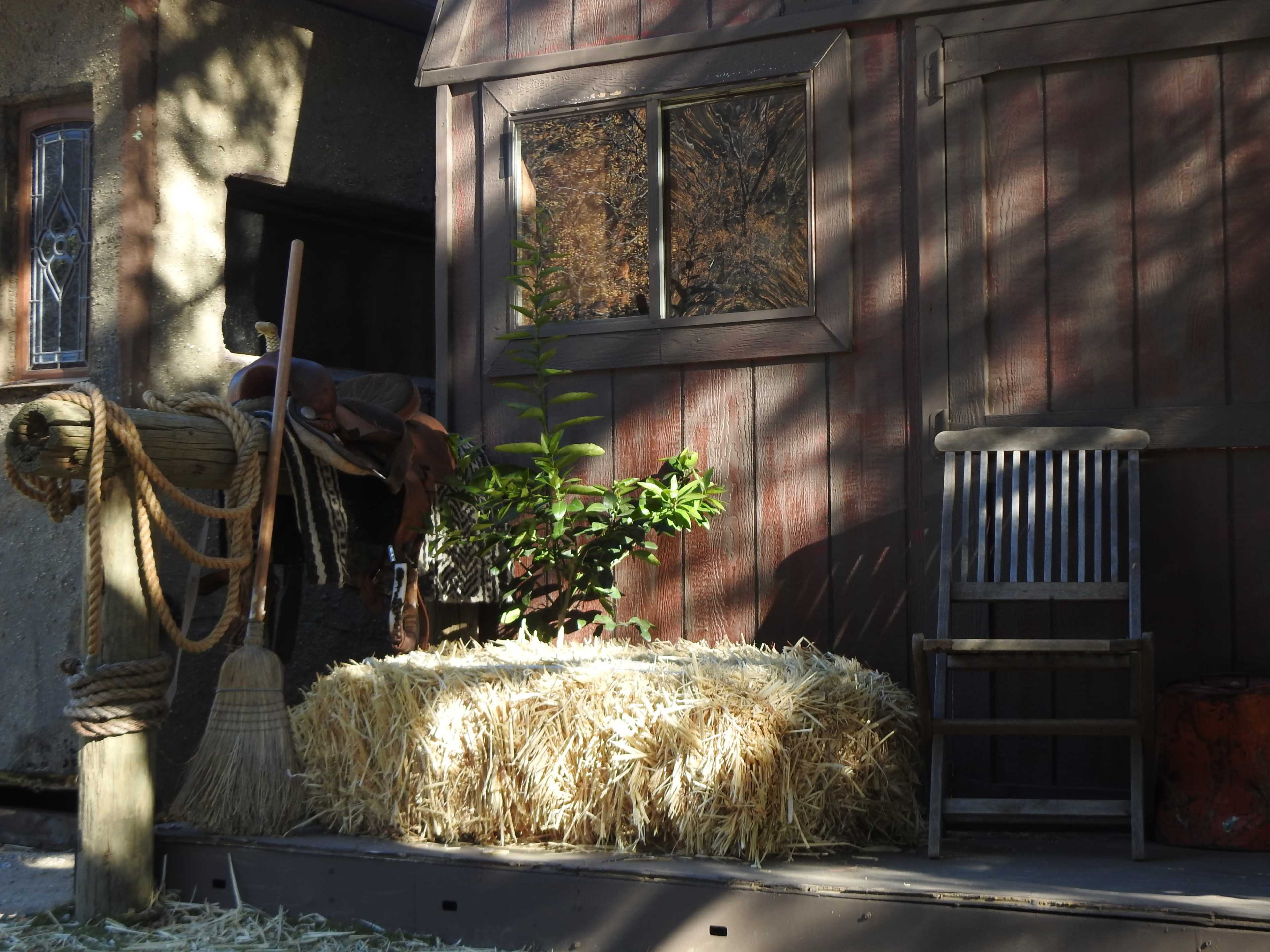 The image shows a rustic scene featuring a straw bale, a wooden chair, and a saddle resting on a post near a barn-like structure.