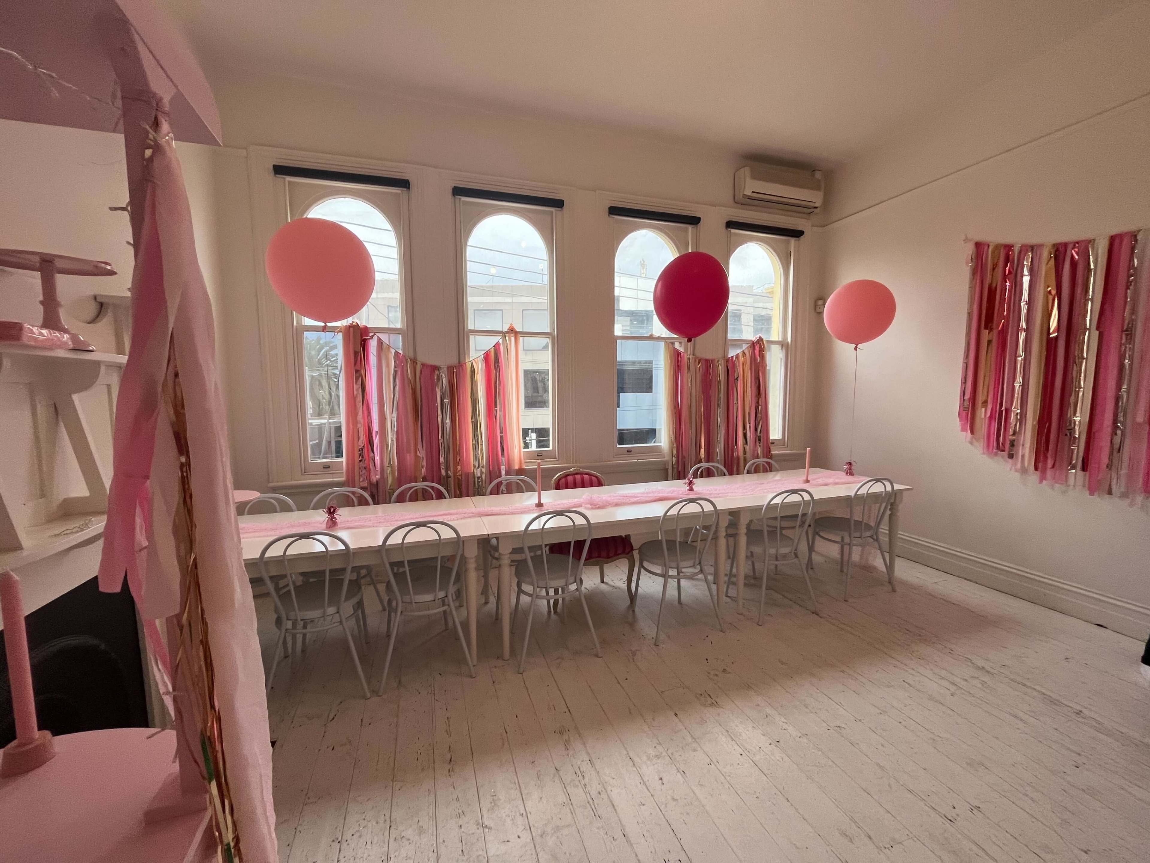 A long white table is set in a room adorned with pink balloons and streamers against a backdrop of large windows.
