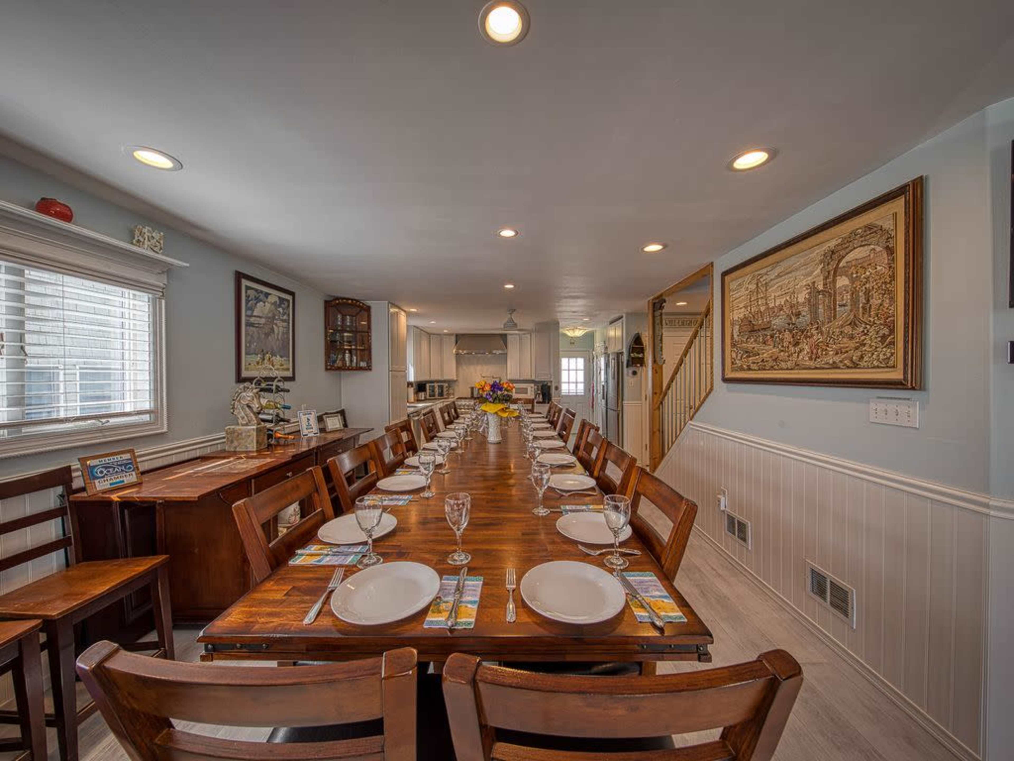 A long wooden dining table is set with plates and glasses in a well-lit dining room that leads to a kitchen and staircase.