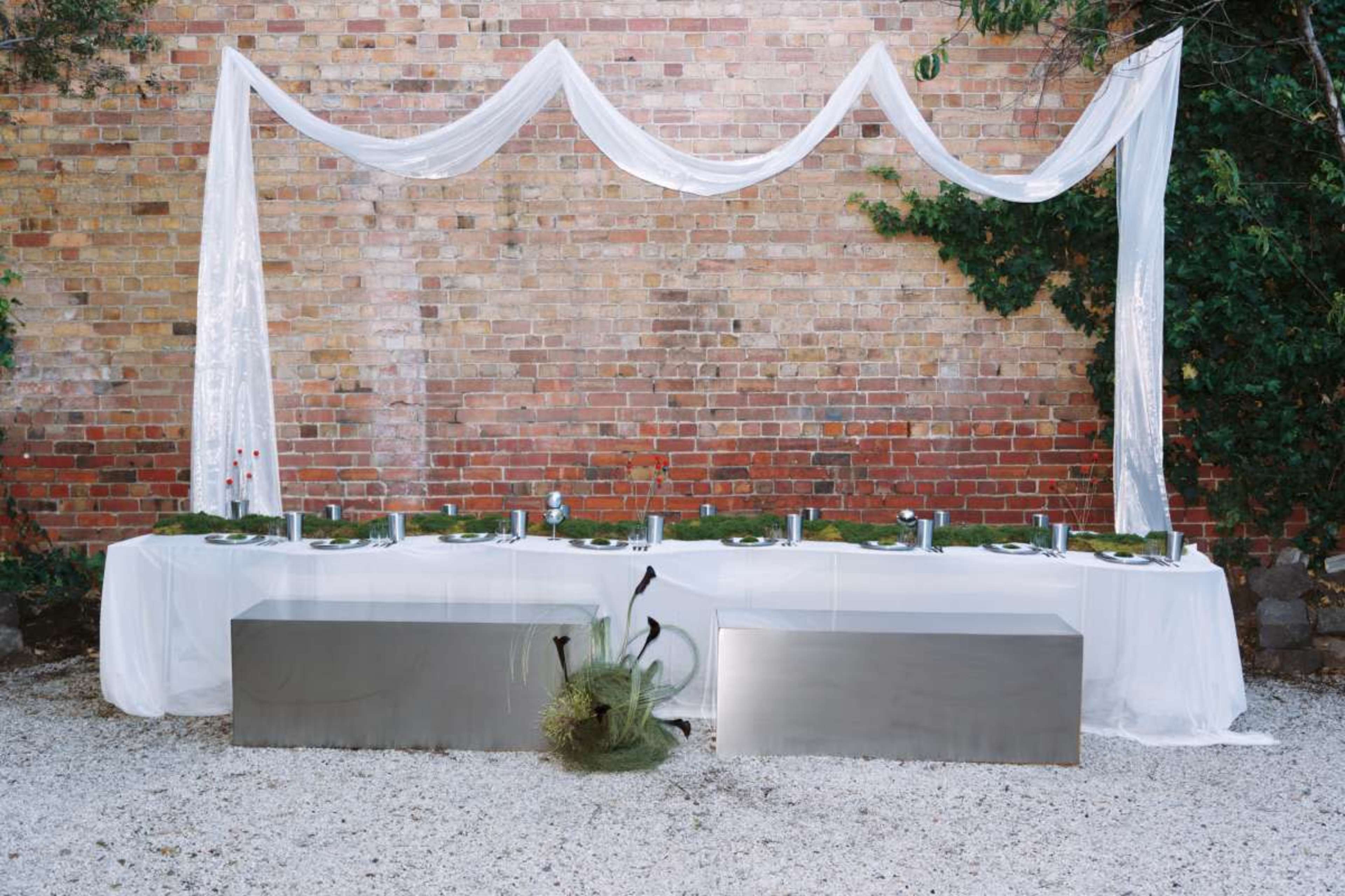 A long table is set outdoors with silver utensils and green accents, positioned against a brick wall and draped with white fabric.