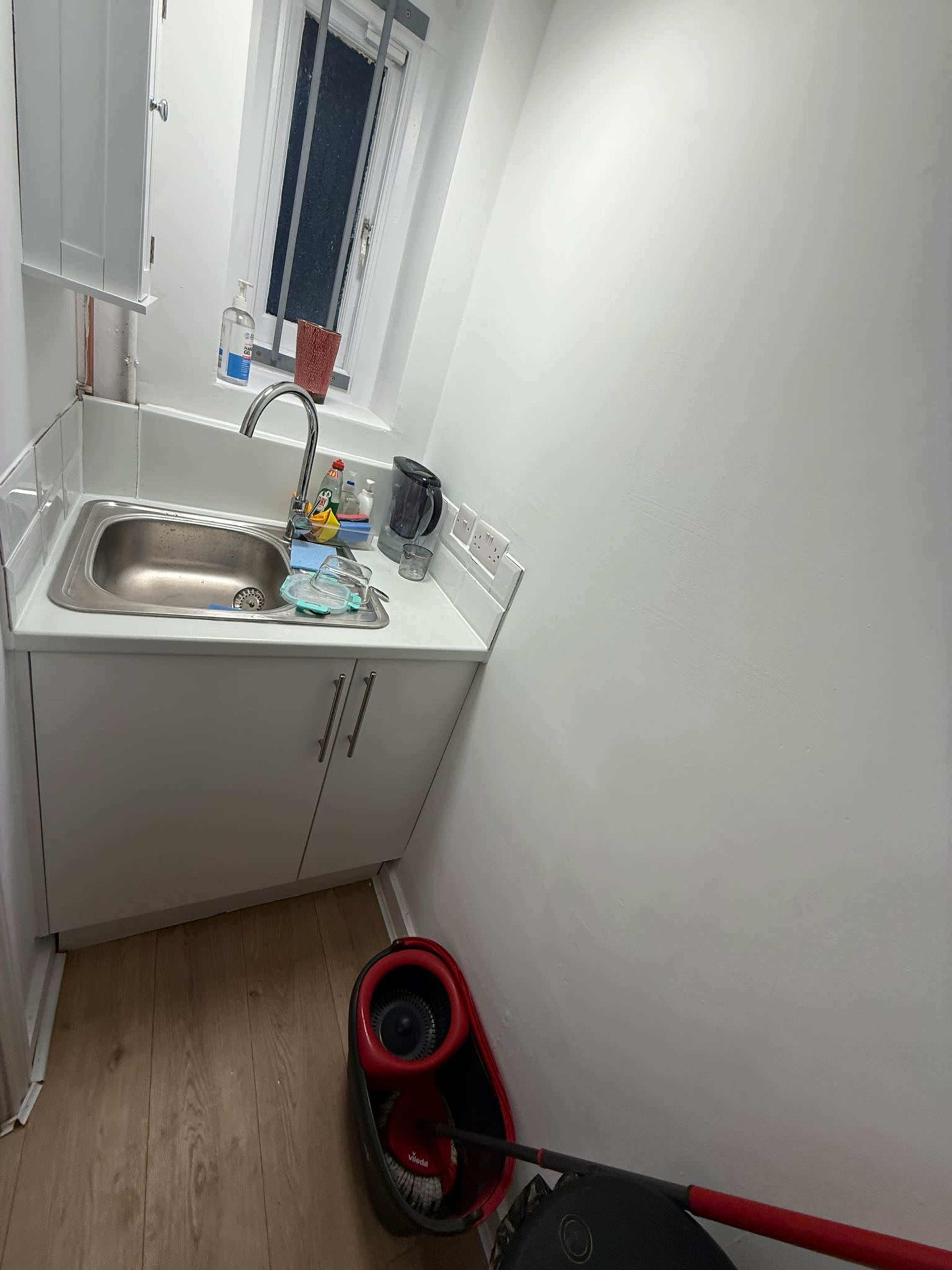The image shows a small kitchen area with a stainless steel sink, a kettle, cleaning supplies, and a vacuum cleaner in the corner.