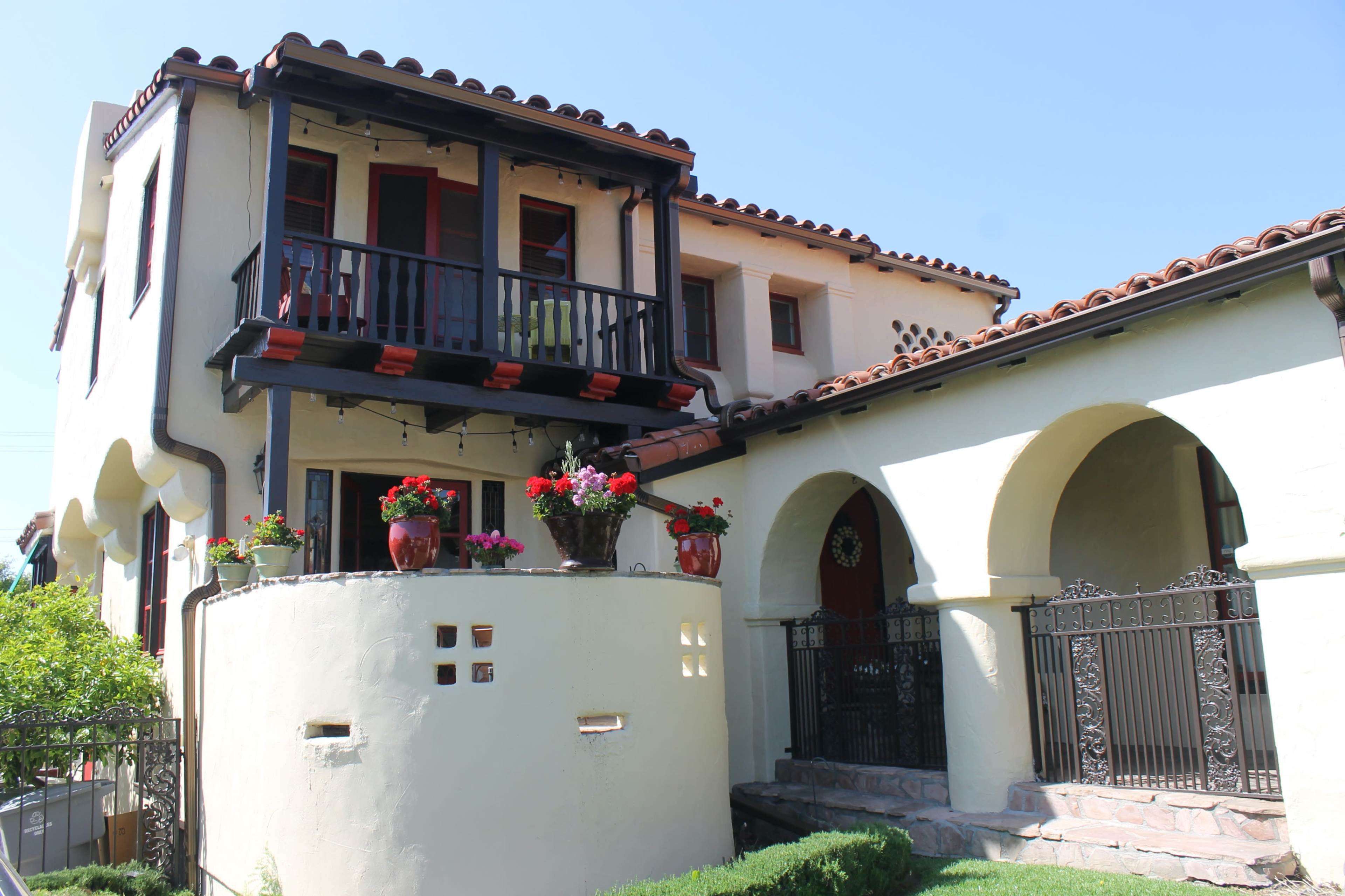 A two-story house with a balcony, decorative flower pots, and a round wall featuring small square windows.