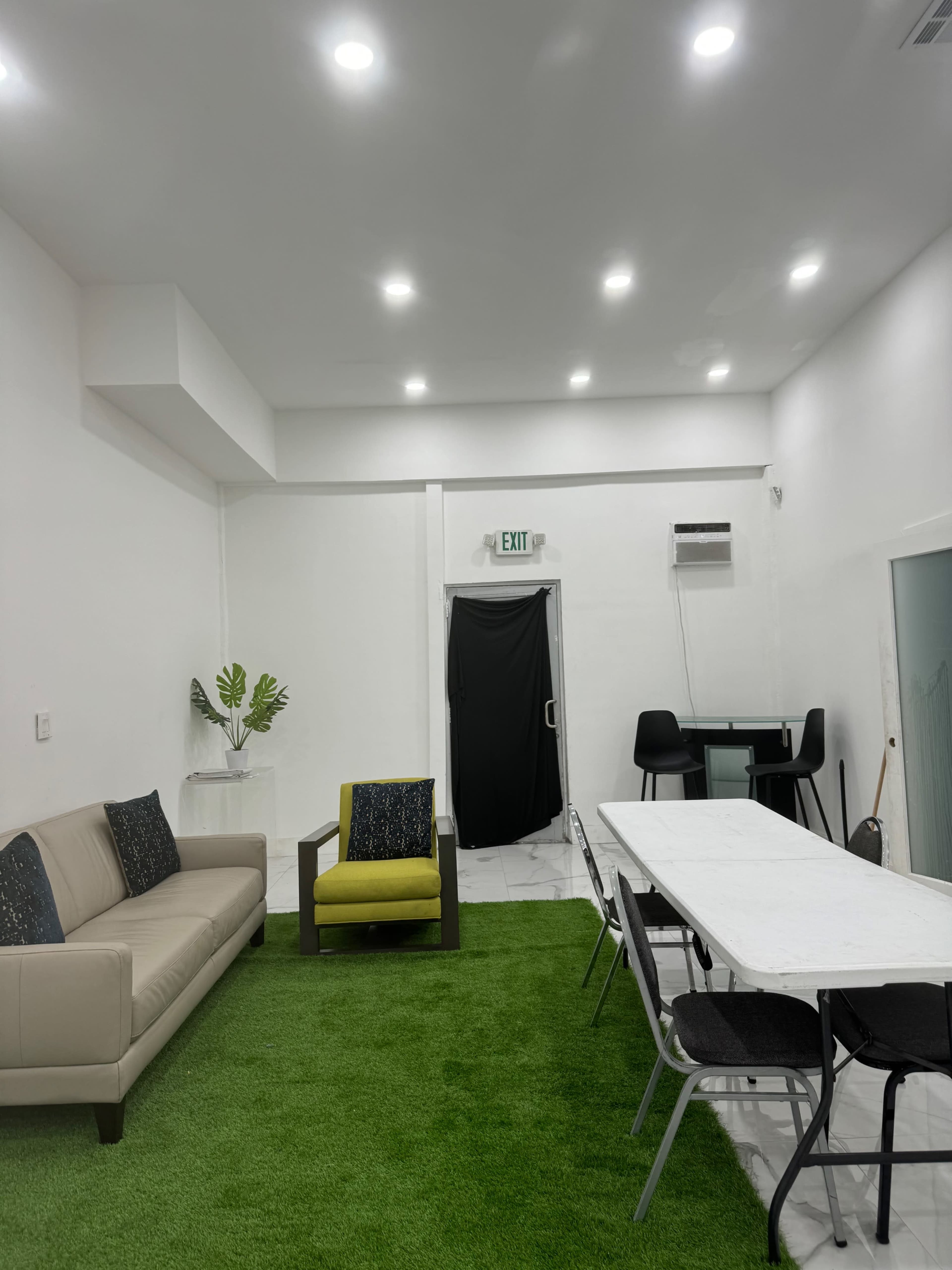 A bright, minimalist room with a sofa, an armchair, a white table with chairs, and green artificial grass flooring.