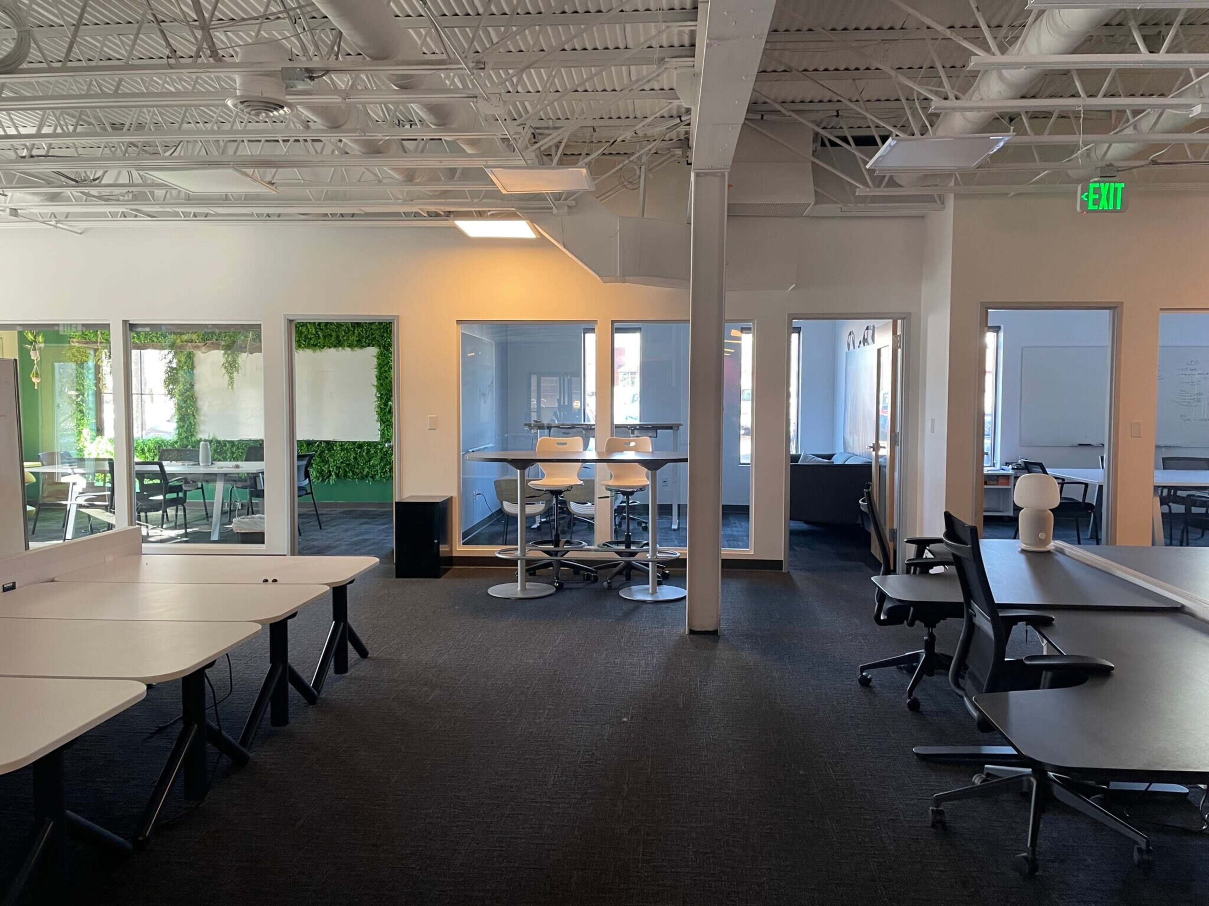 The image shows a modern office space with multiple workstations, a meeting area with high tables, and glass walls providing a view of a green accent wall.