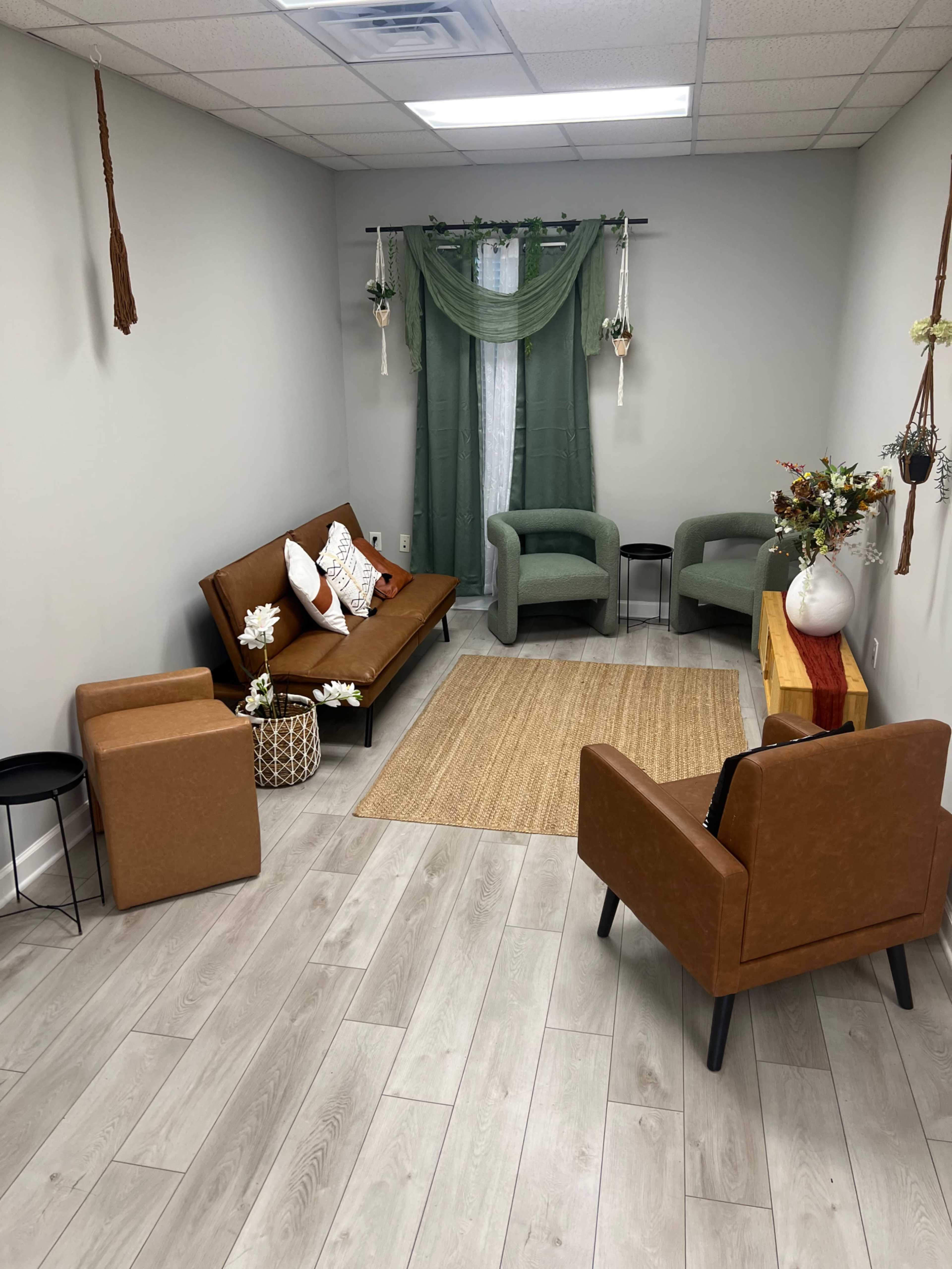 The image shows a cozy waiting area furnished with a brown sofa, two green armchairs, a wooden side table, and decorative plants, all set on light wood flooring.