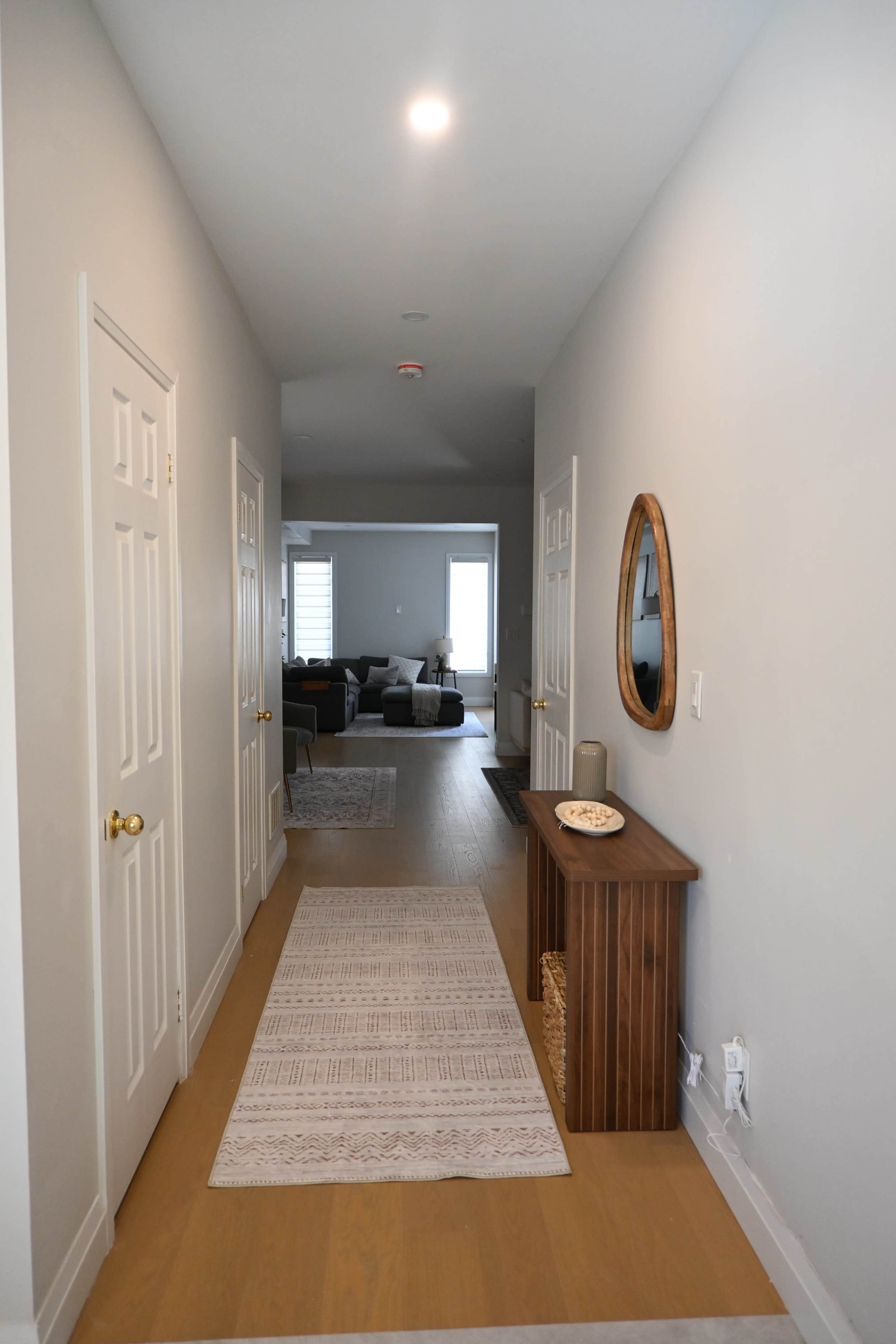 The image shows a long, narrow hallway with light gray walls, a rug on the floor, a wooden console table, and a circular mirror.