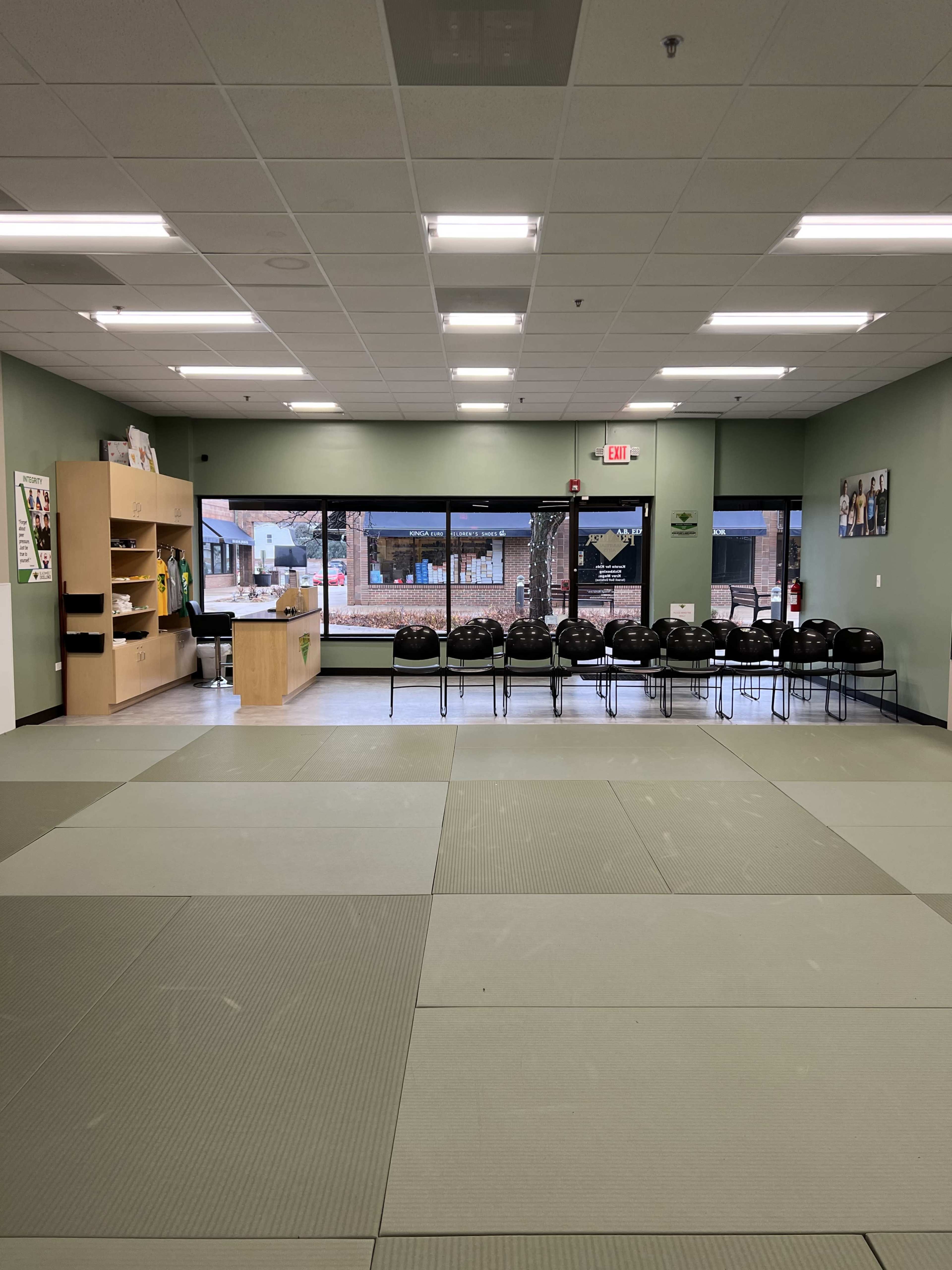 The image shows a dojo with a mat area in the foreground and a row of black chairs lined up against a wall, along with a reception area and large windows in the background.