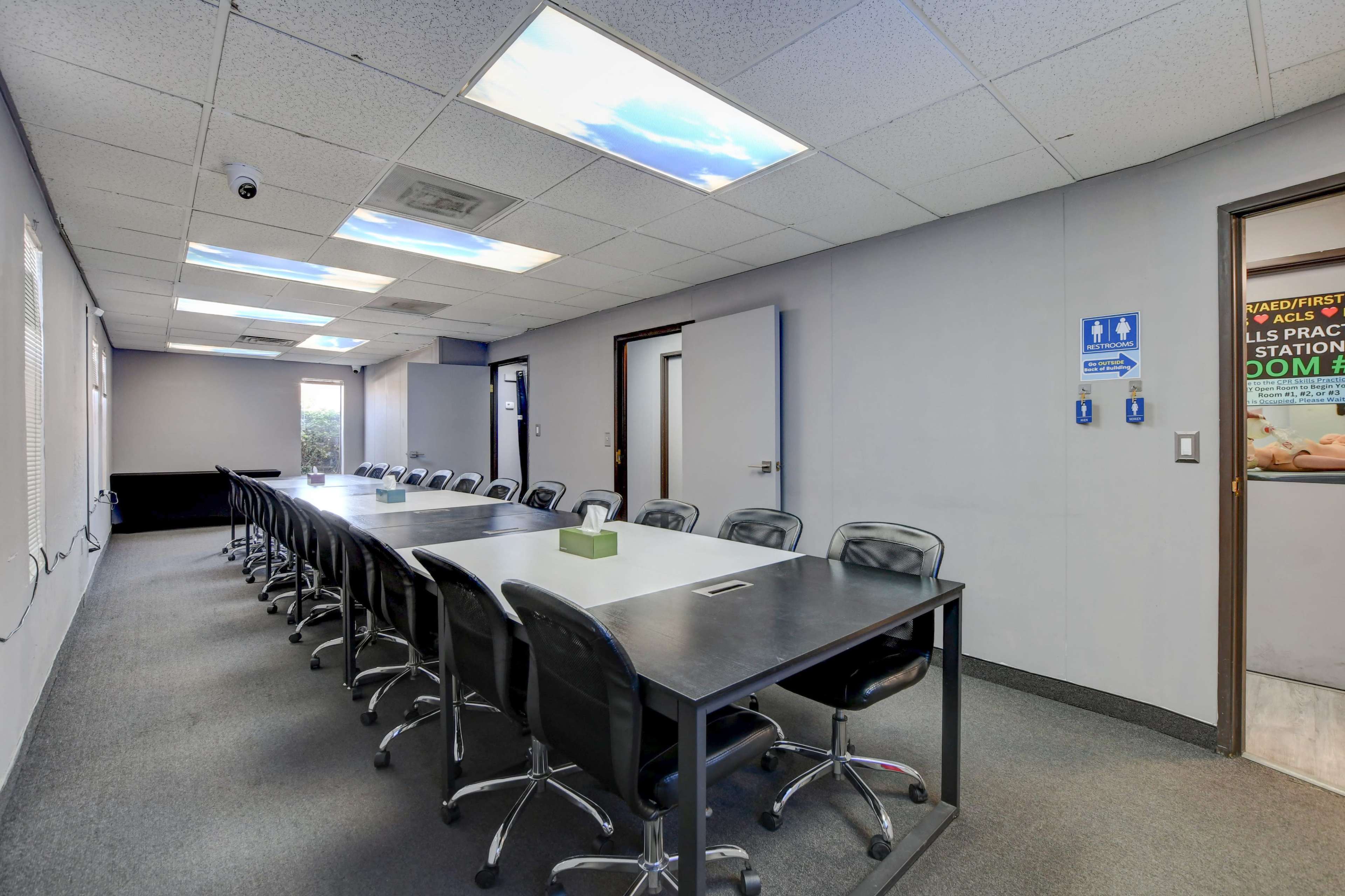 A long conference table with black chairs is positioned in a brightly lit meeting room, featuring windows that provide natural light and a door to another space.