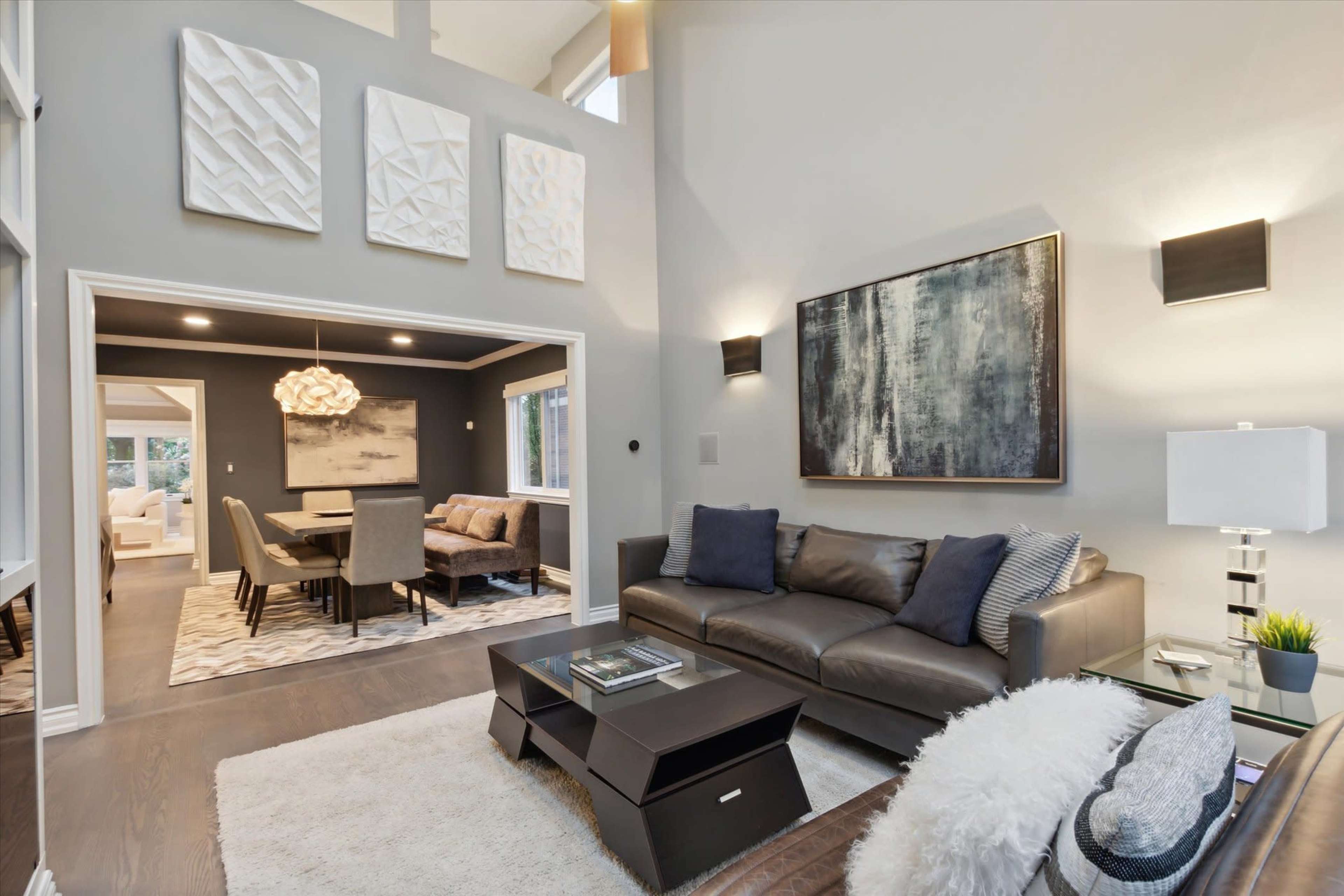A modern living room features a dark leather sofa facing a sleek coffee table, with large artwork on the walls and a dining area visible through an archway.