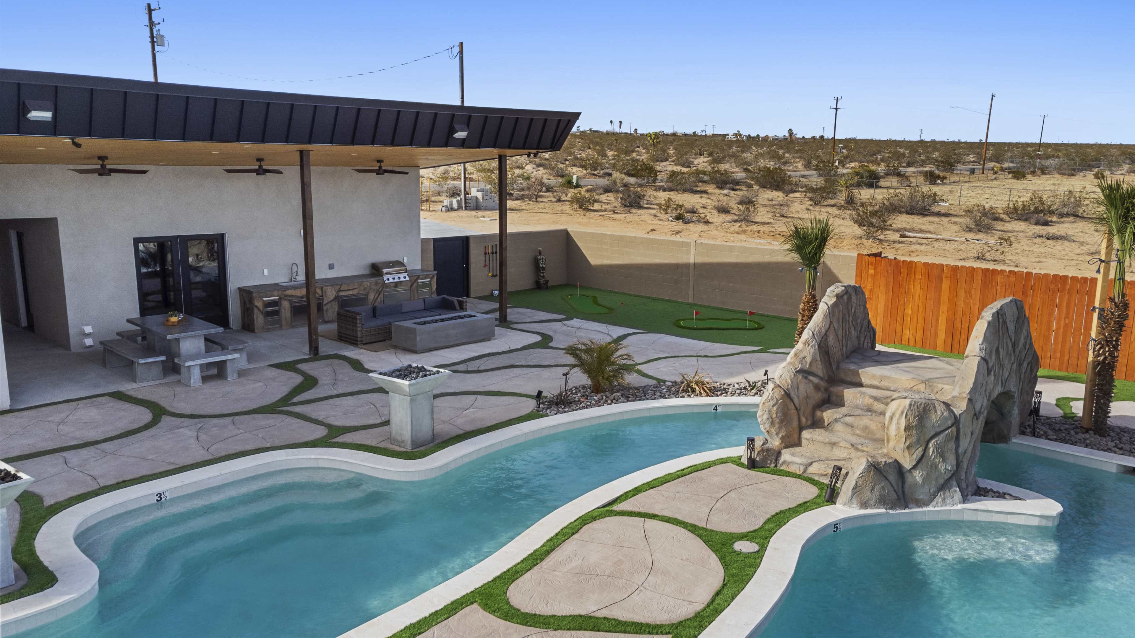 A modern backyard with a swimming pool featuring a rock formation waterfall, surrounded by concrete pathways and an outdoor seating area.