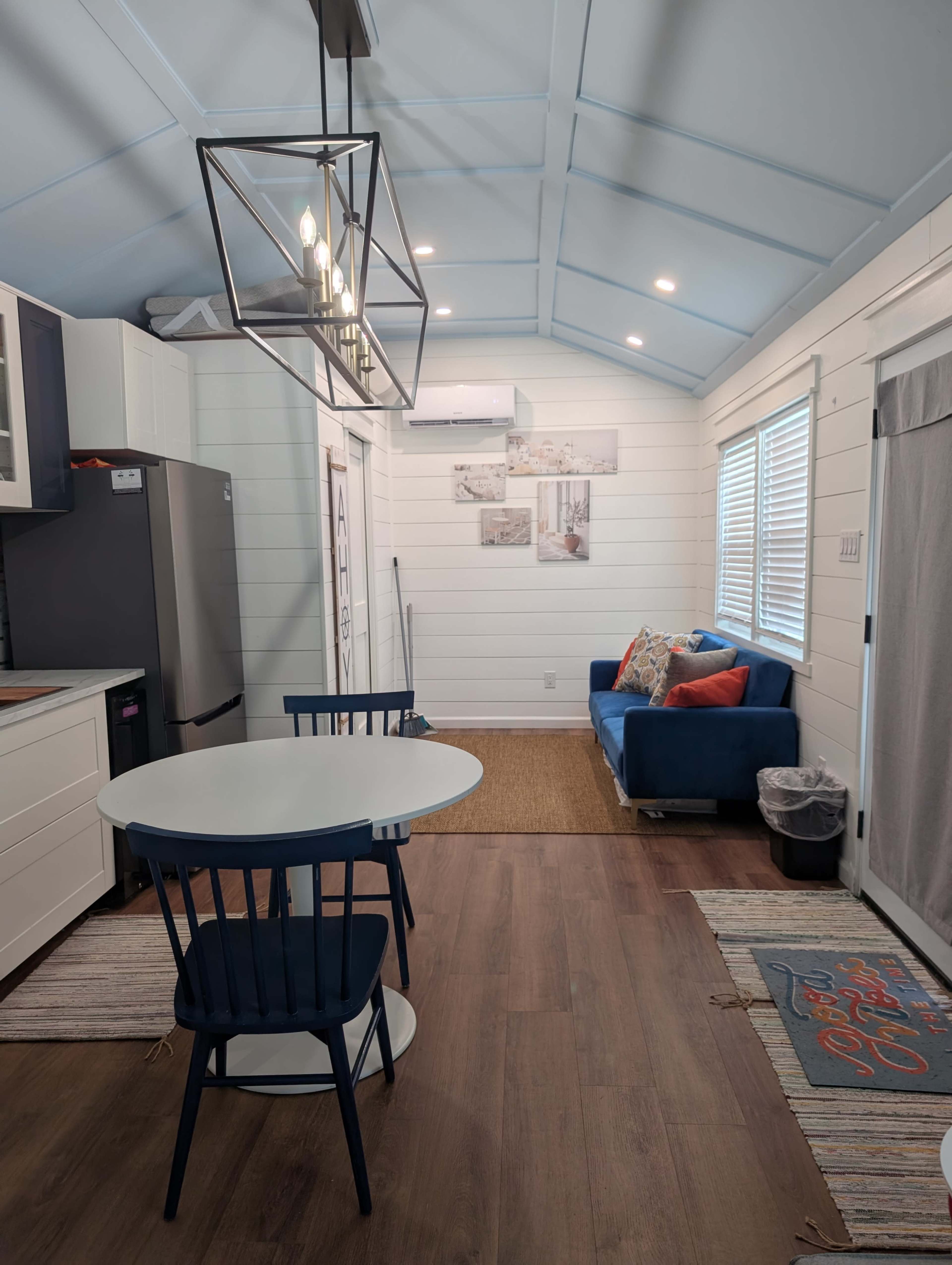The image shows a small, well-lit interior of a cabin or tiny house with a dining table and chairs, a sofa, and modern kitchen appliances.