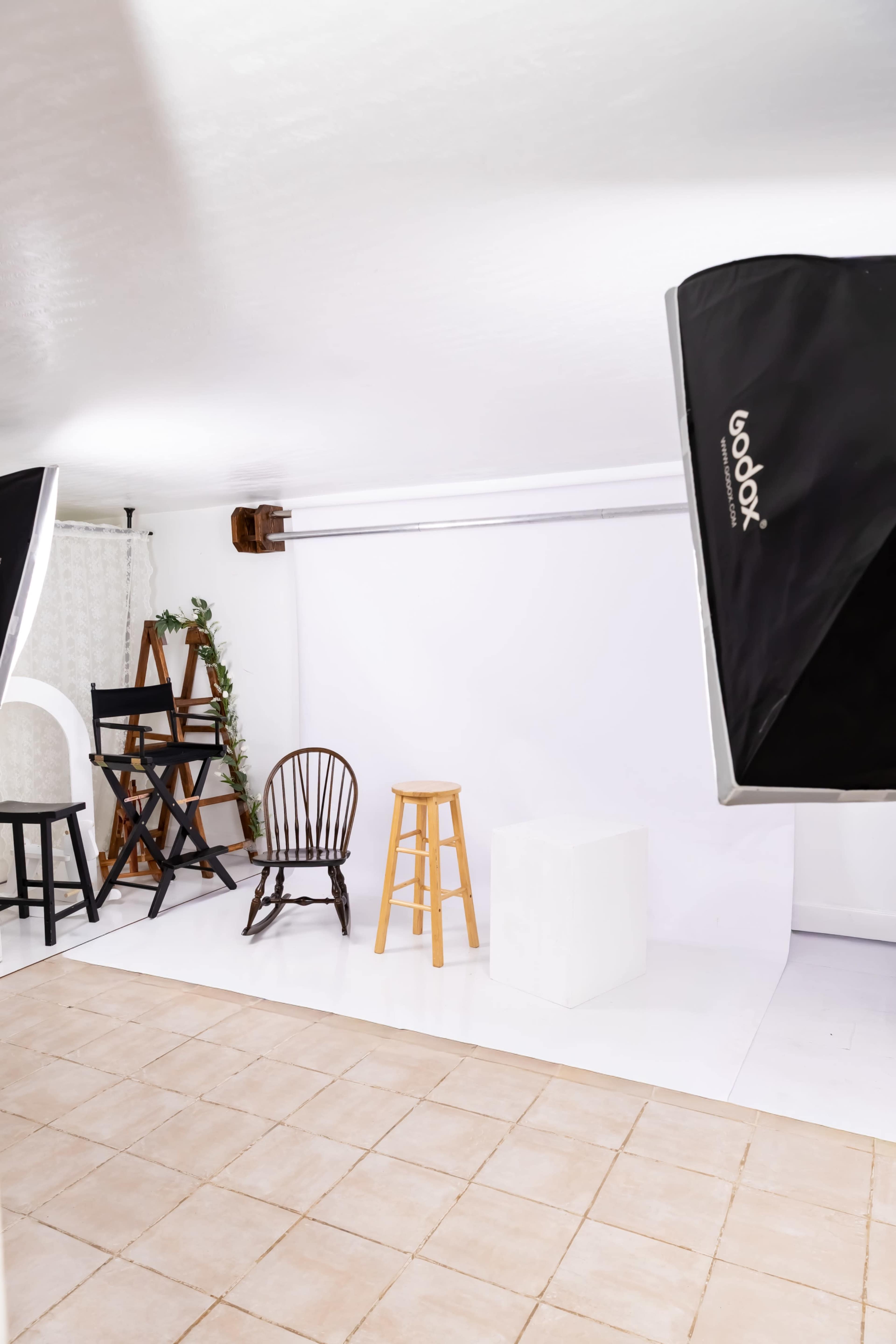 The image shows a photography studio with a white backdrop and various chairs and stools arranged on a tiled floor.