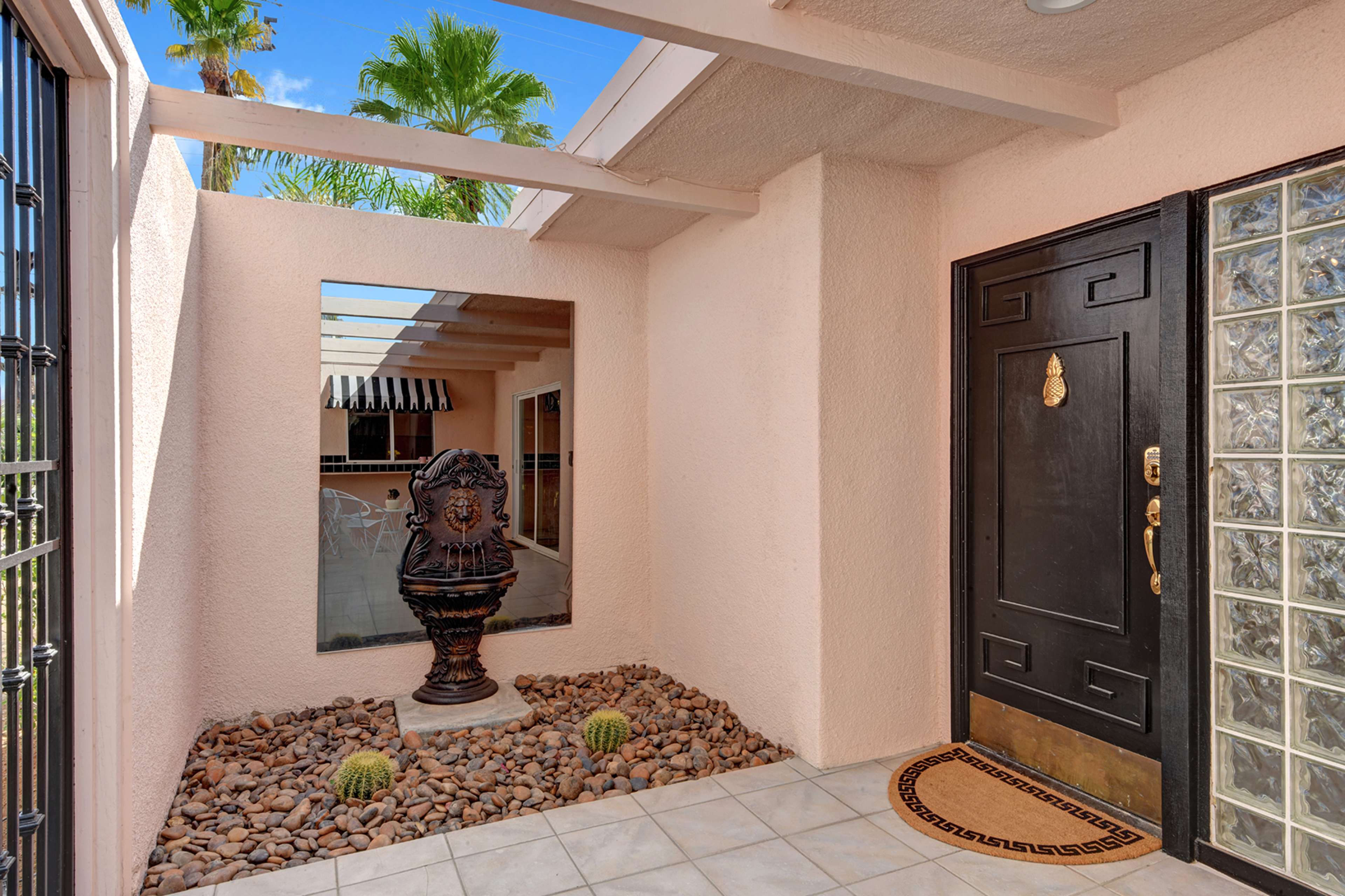 The image shows an entrance with a large black door, a decorative fountain in a landscaped area with pebbles, and a reflective mirror on the wall.